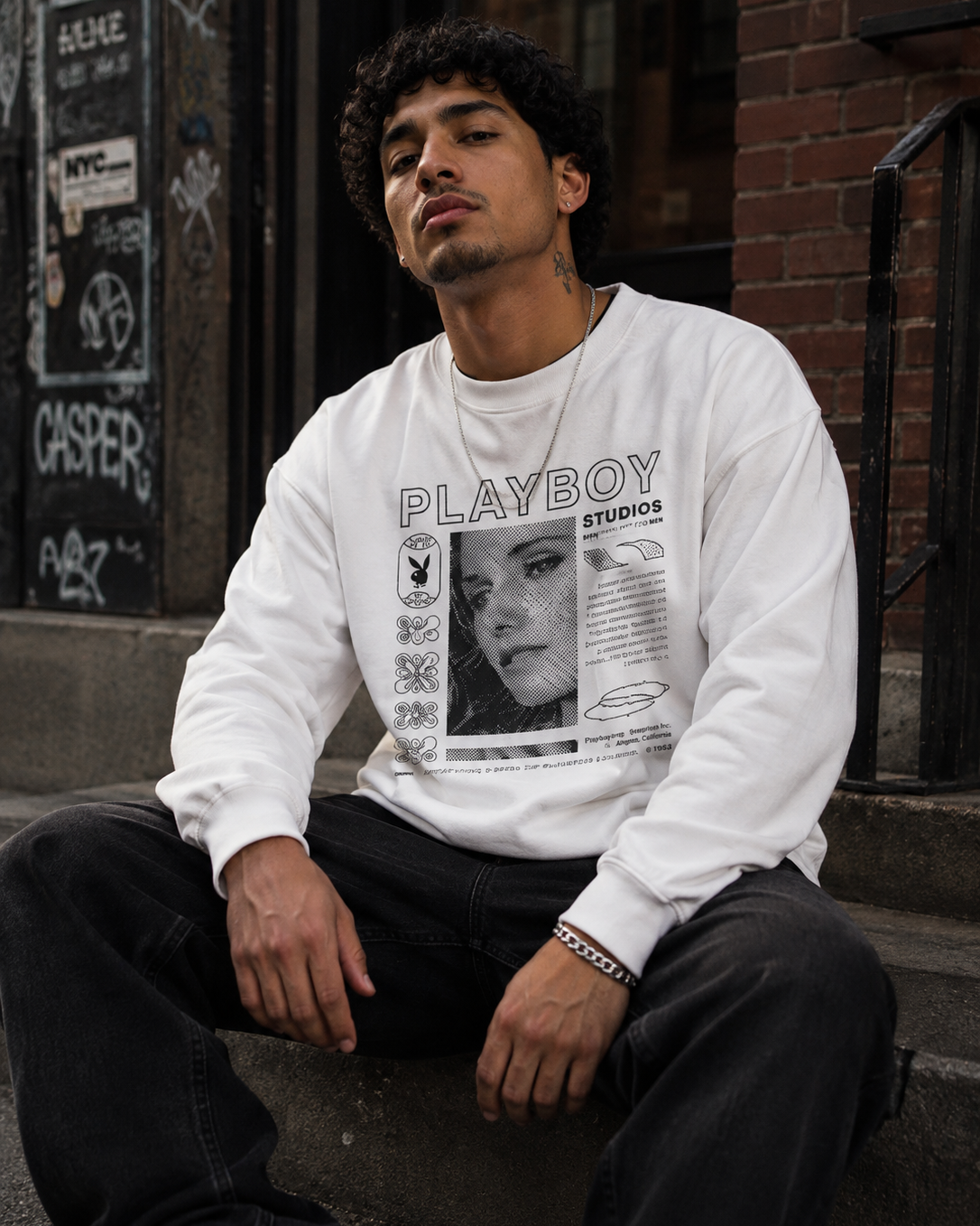 A man sitting on outdoor concrete steps in front of a brick building, wearing a white long-sleeve crewneck sweatshirt with a black and white Playboy Studios graphic print on the front, paired with black jeans. #Streetwearmen