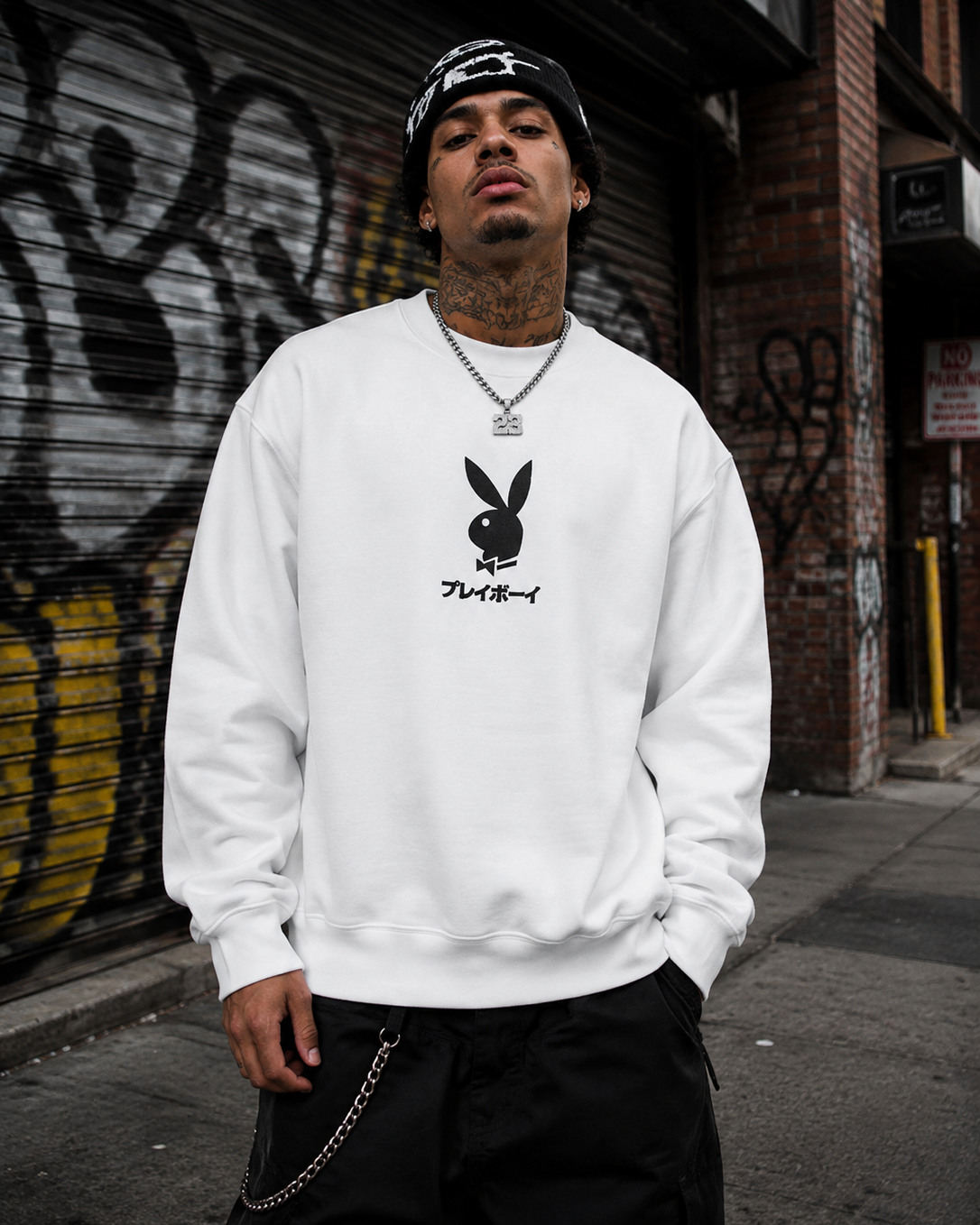 A man wearing a white crewneck sweatshirt with a black Playboy bunny and script logo on the chest, paired with a black beanie, standing in front of a graffiti-covered metal shutter. #Streetwearmen