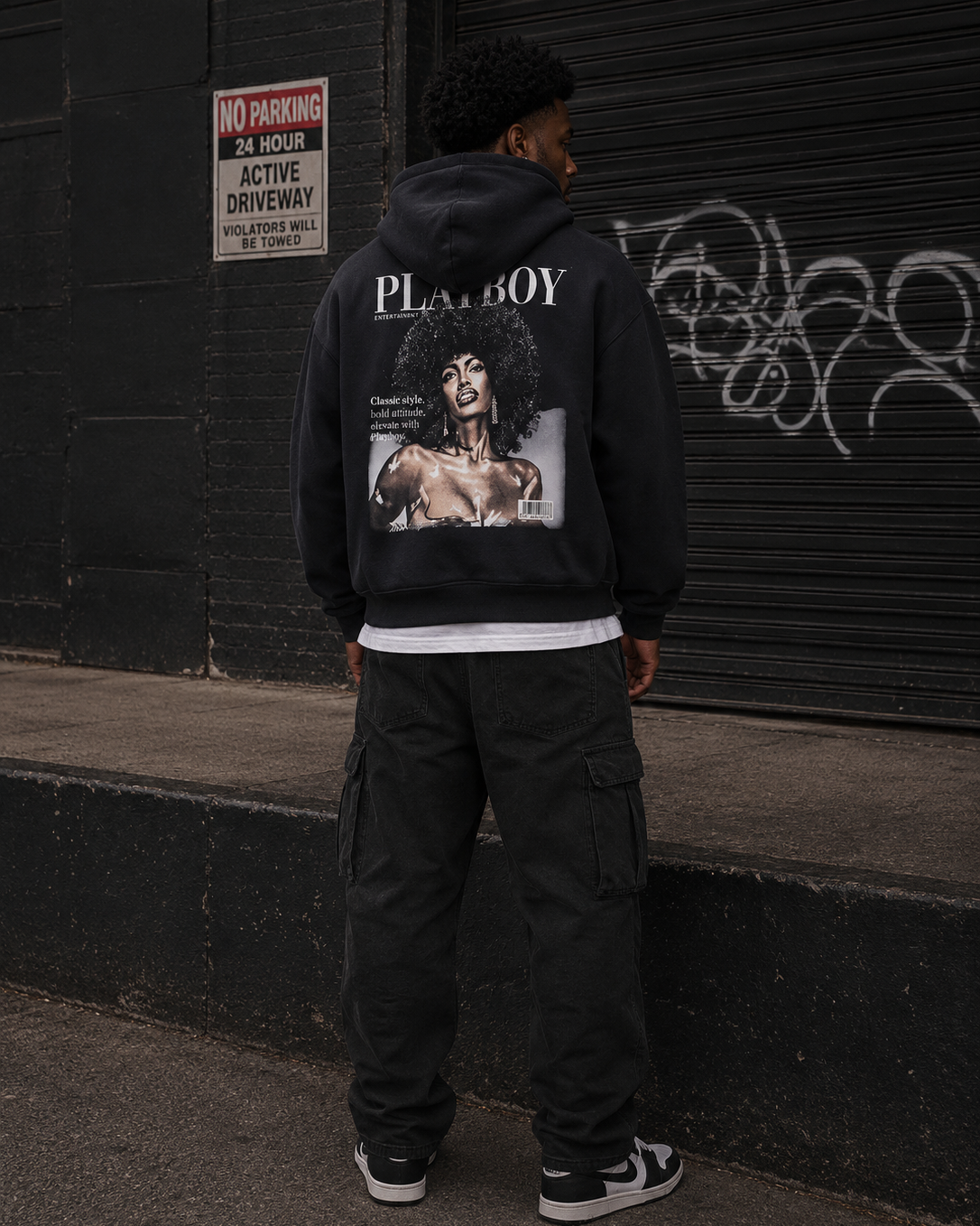 A man stands with his back to the camera wearing a black Playboy hoodie with a large graphic print on the back, paired with black cargo pants and black-and-white sneakers, set against a dark urban wall with graffiti. #Streetwearmen