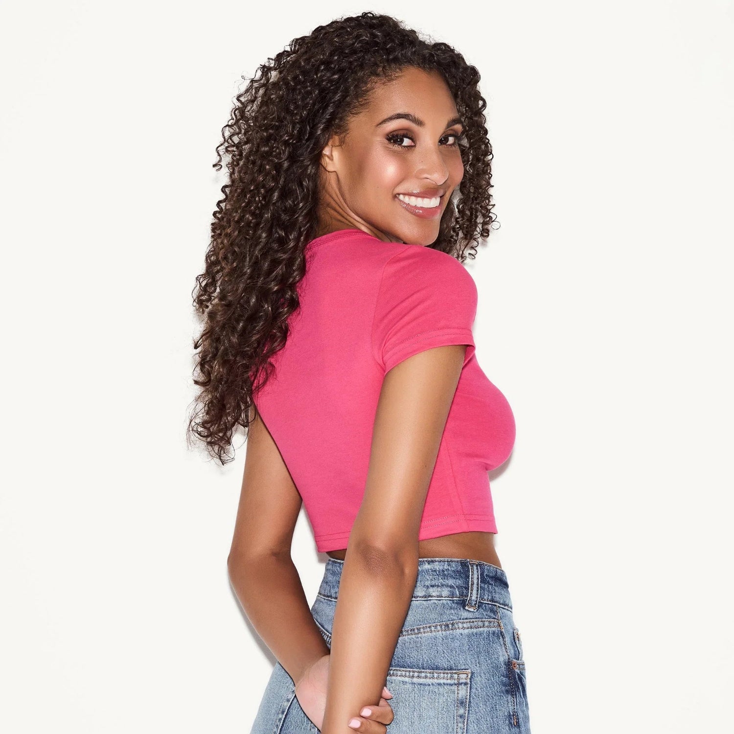 Woman wearing a pink crop baby tee and high-waisted blue jeans, smiling over shoulder