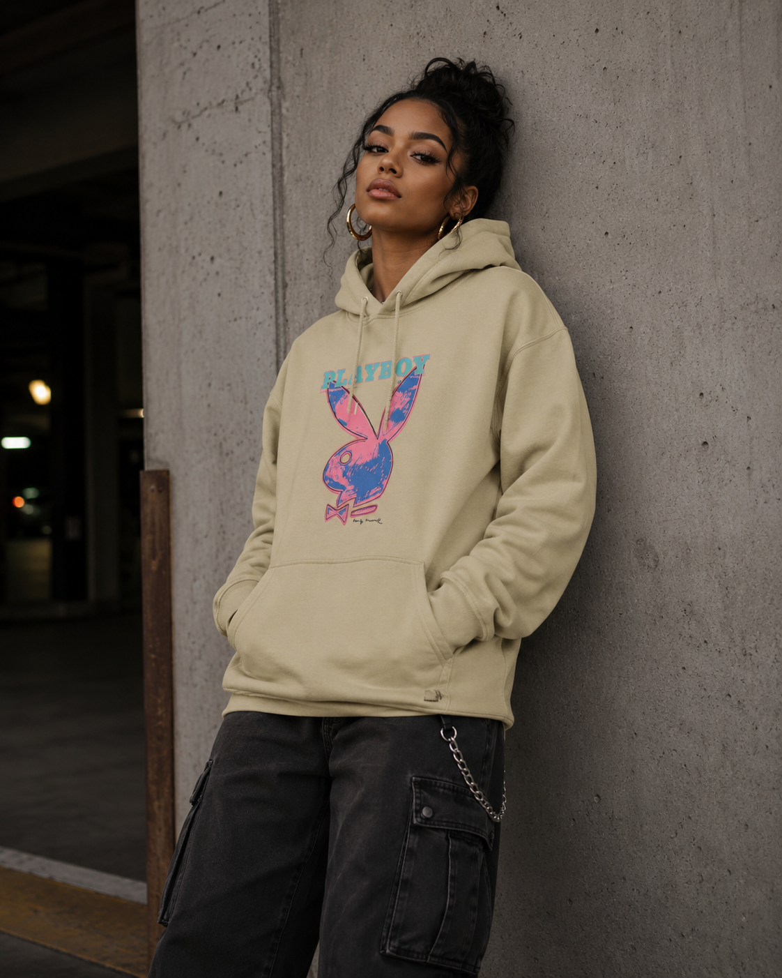 A woman wearing a beige hoodie with a colorful graphic print and dark cargo pants, leaning against a concrete wall. #Streetwearwomen