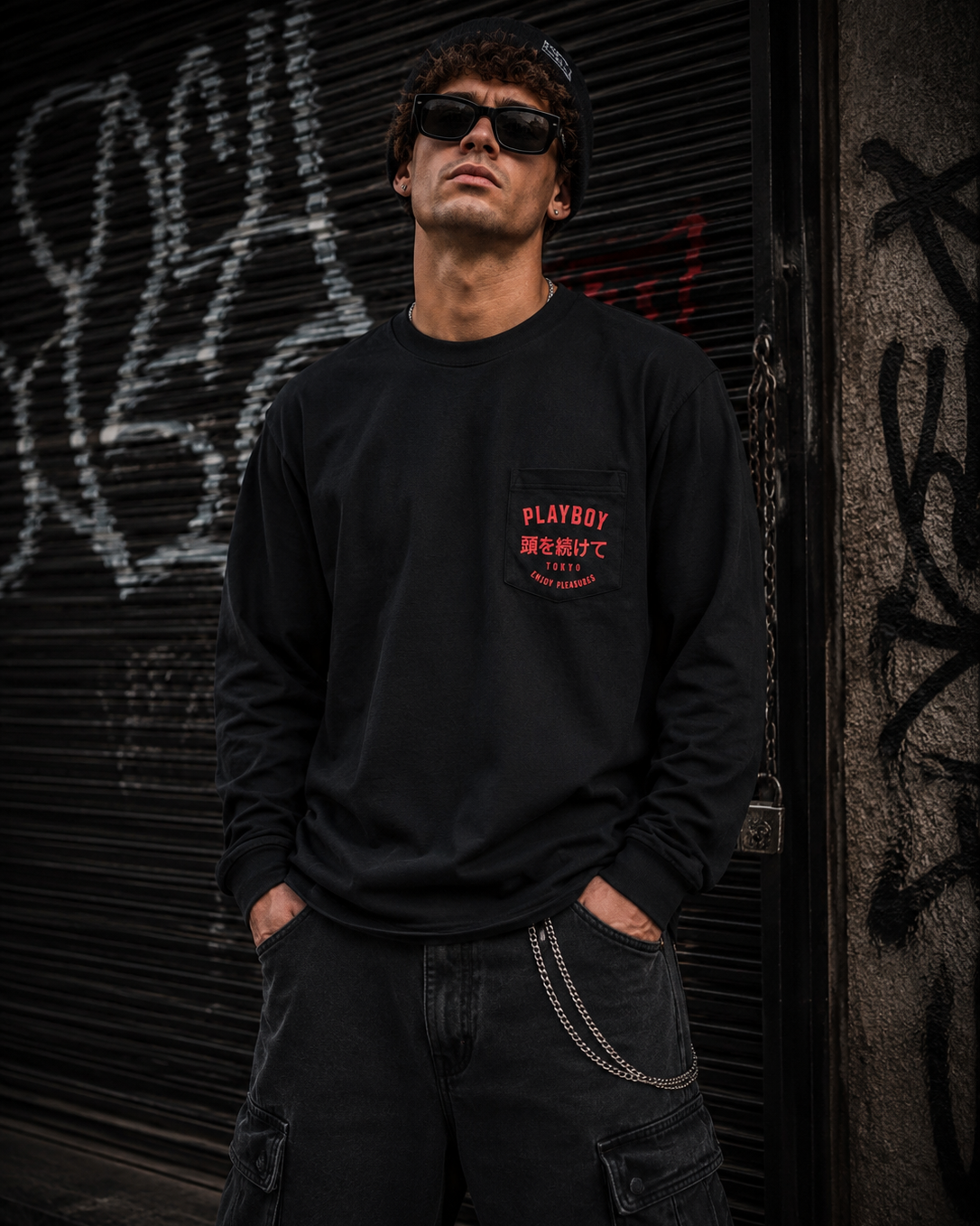 A man wearing a black long-sleeve crewneck sweatshirt with a red 'PLAYBOY' chest graphic and sunglasses, standing in front of a graffiti-covered metal shutter. #Streetwearmen