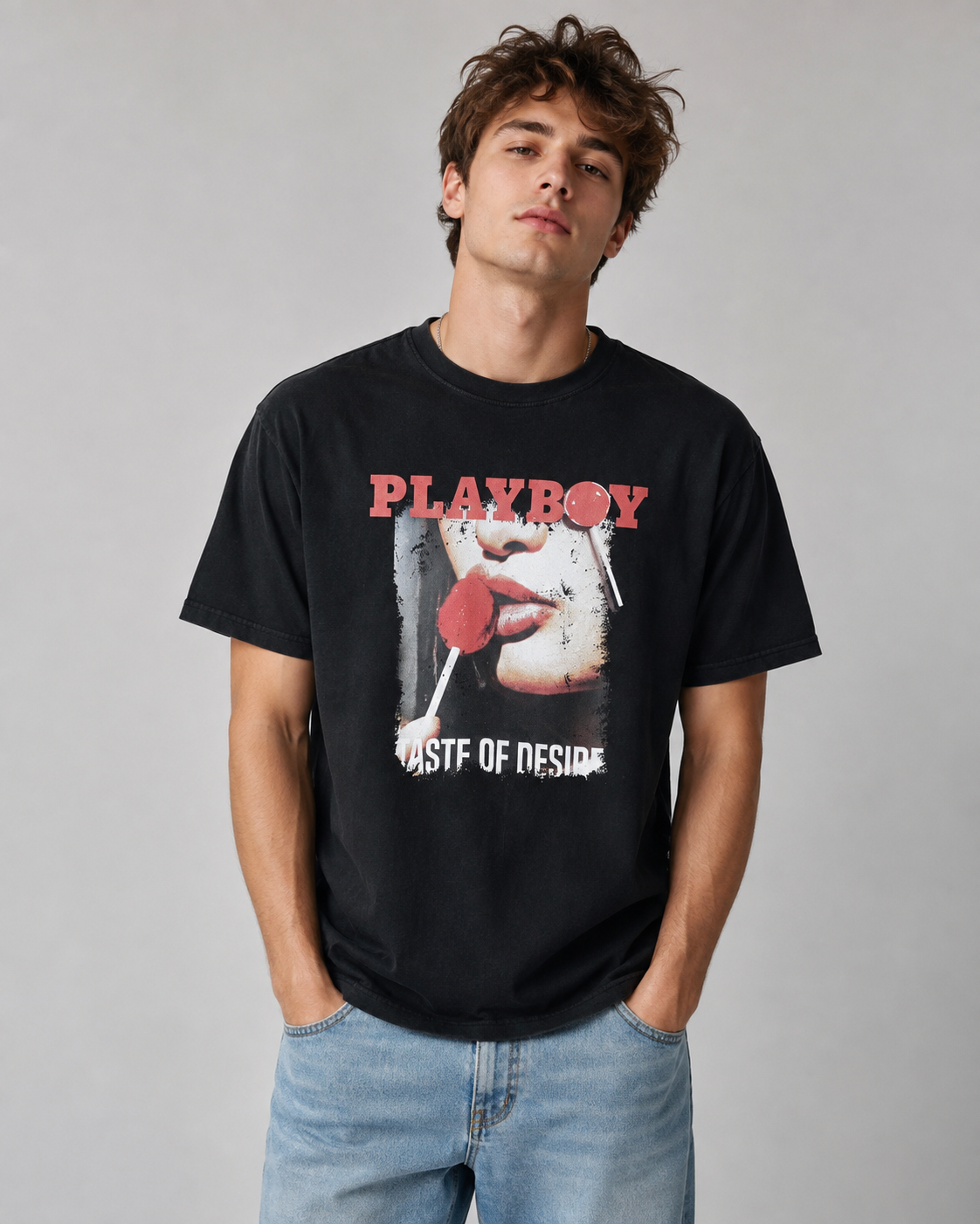 A man wearing a black graphic t-shirt with a red and white Playboy 'Most of These' print, paired with light blue jeans, standing against a neutral gray background.