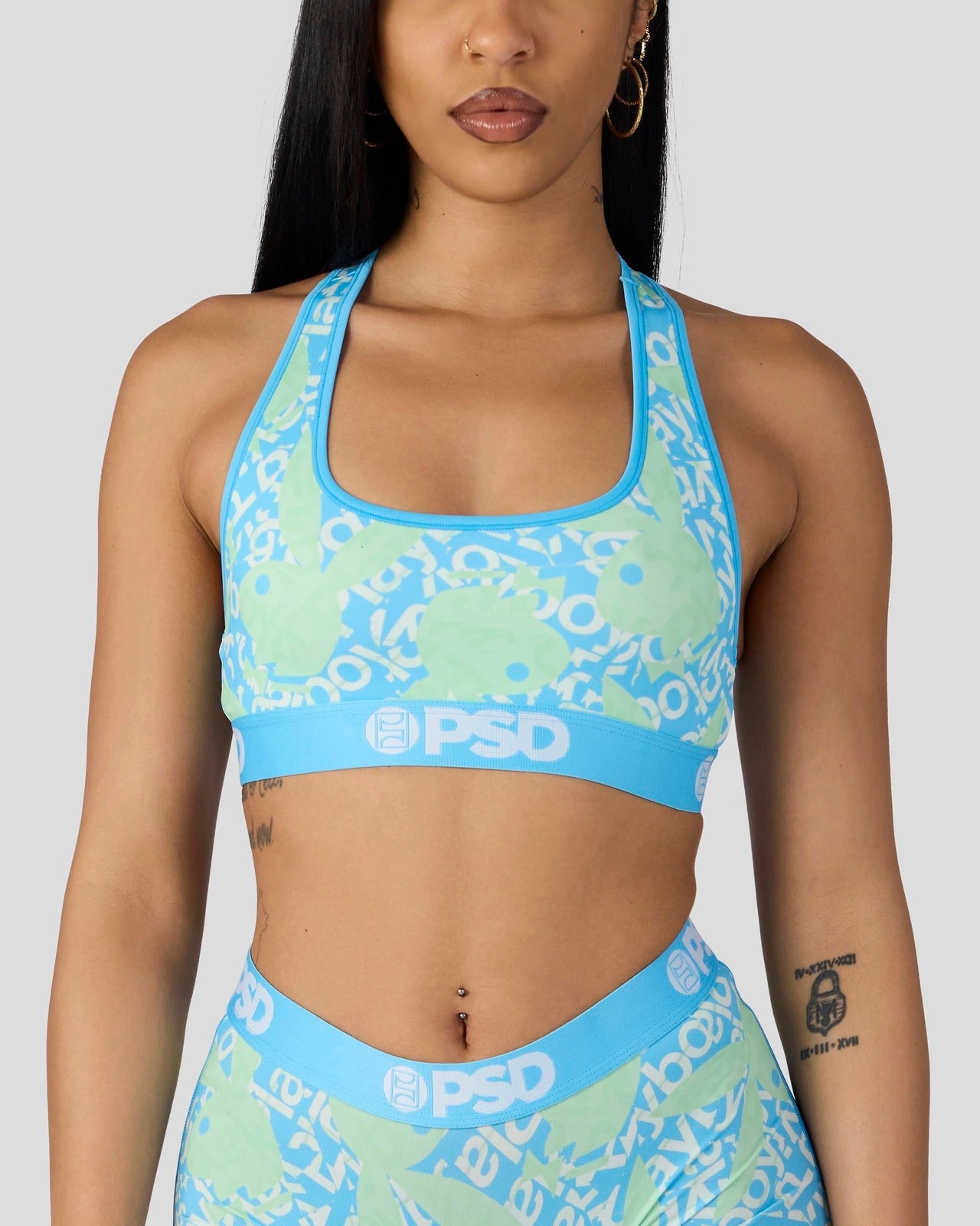 A woman wearing a matching light blue and mint green sports bra and high-waisted leggings set with a white 'PSD' logo print, shown against a solid gray background.