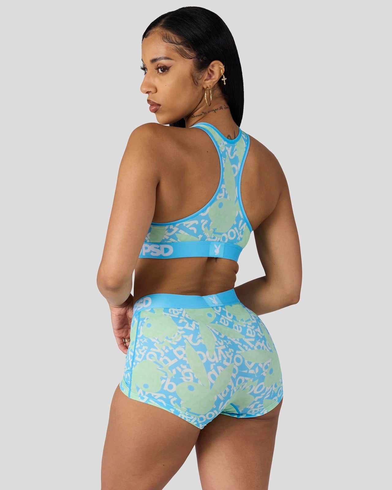 A woman shown from the back wearing a matching two-piece activewear set with a light blue and mint green abstract pattern. The set includes a sports bra and high-waisted shorts, photographed against a solid light gray background.