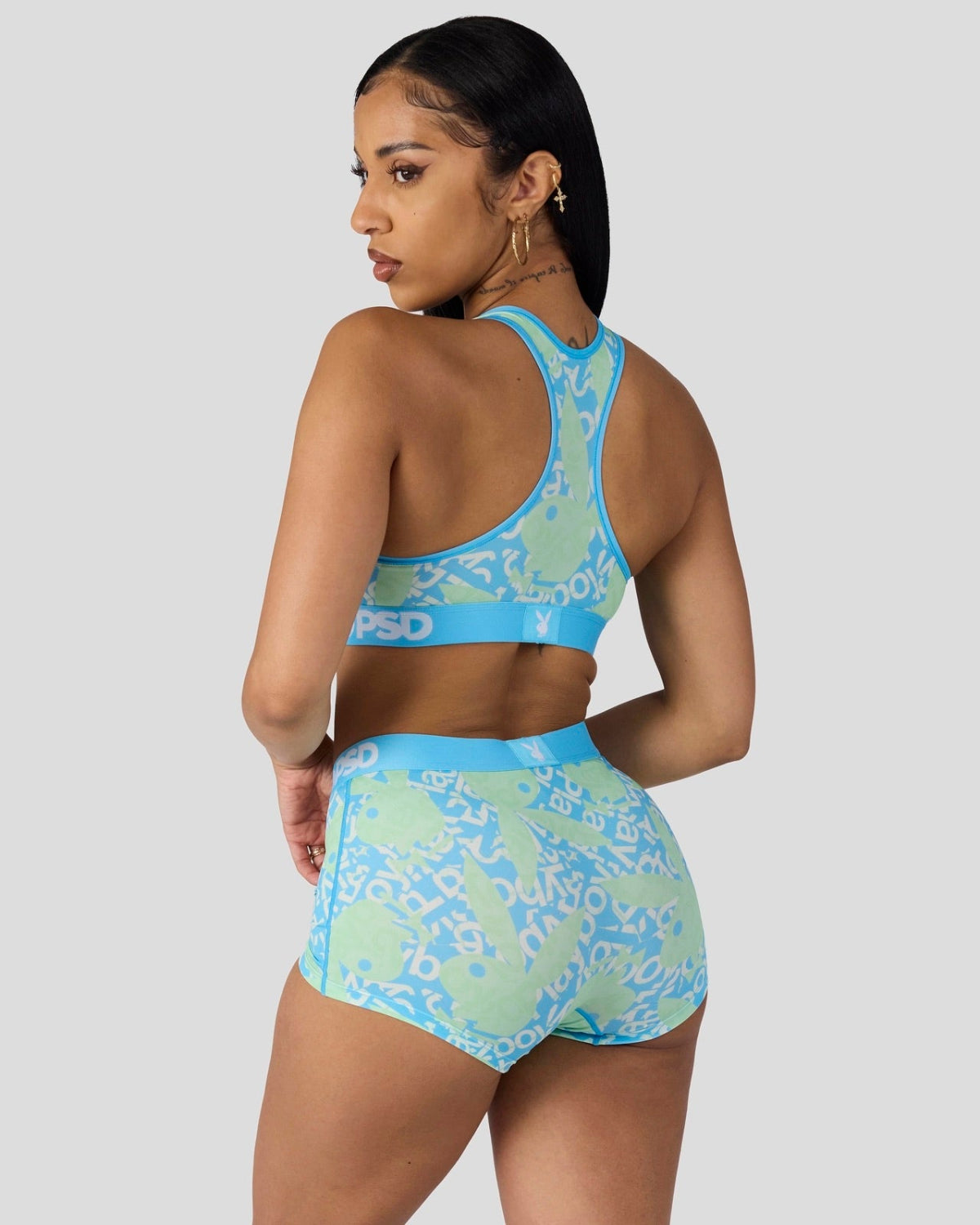 A woman shown from the back wearing a matching two-piece activewear set with a light blue and mint green abstract pattern. The set includes a sports bra and high-waisted shorts, photographed against a solid light gray background.