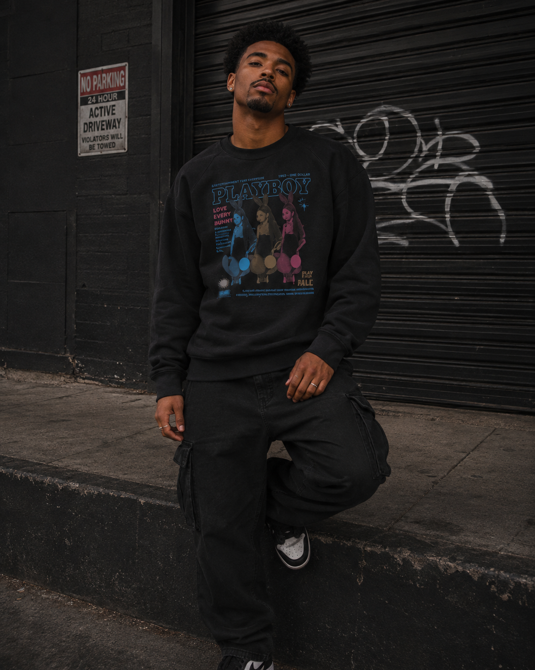 A man wearing a black long-sleeve crewneck sweatshirt with a colorful graphic print on the front, paired with black cargo pants and white sneakers, posing on concrete steps in front of a black metal shutter. #Streetwearmen