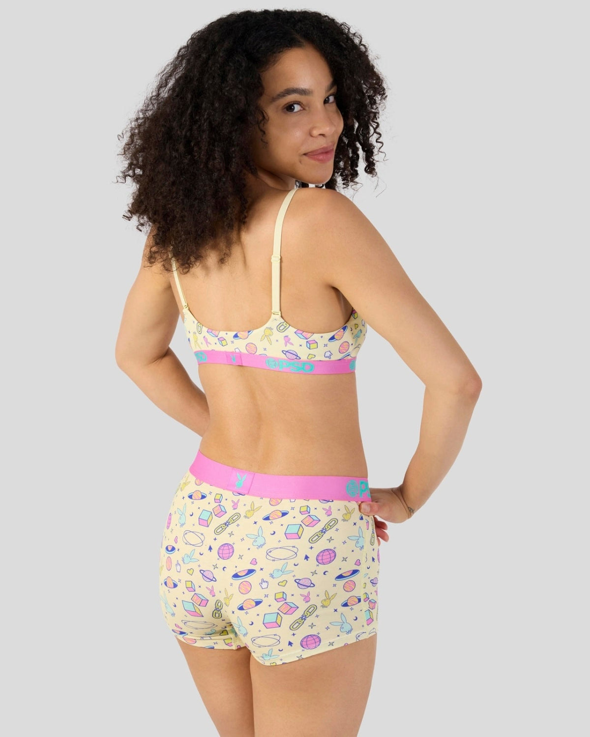 A woman modeling a matching two-piece lingerie set with a colorful candy print on a pale yellow background, featuring a pink waistband and a pink band on the bra.