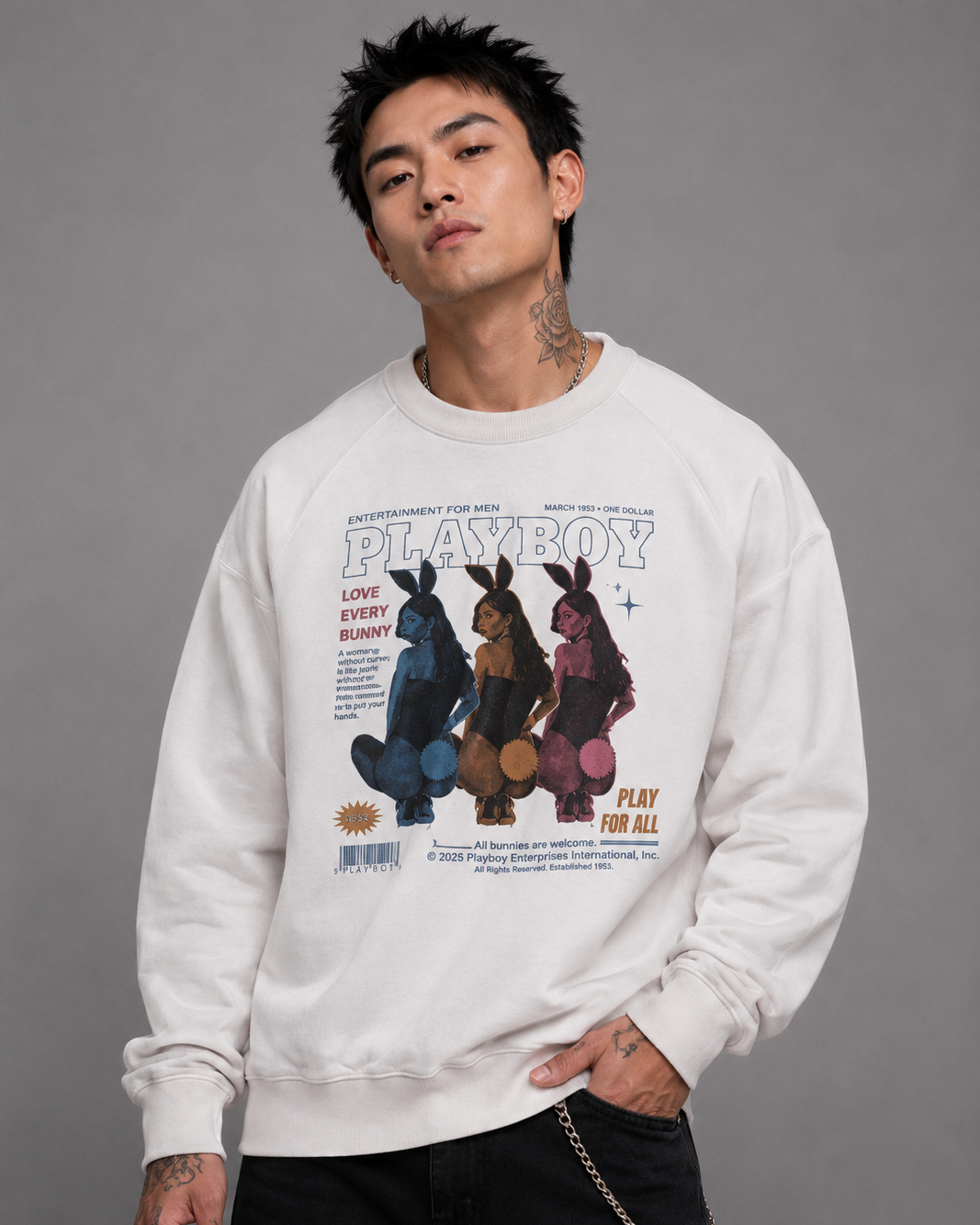 A man wearing a white Playboy crewneck sweatshirt with a vintage-style bunny graphic on the front, paired with black pants, against a solid gray studio background.