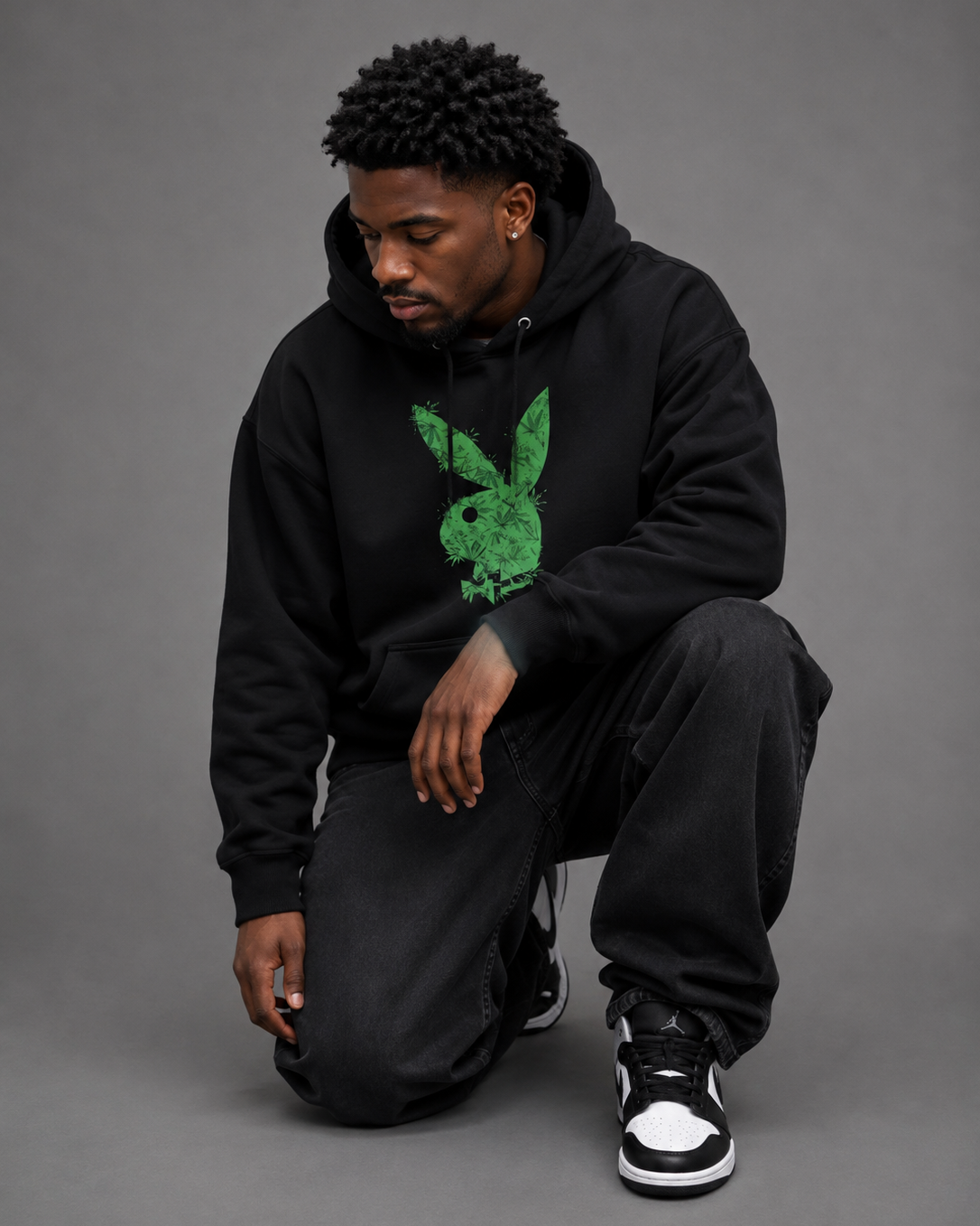A man wearing a black hoodie with a green distressed Playboy bunny graphic, black jogger pants, and black and white sneakers, kneeling against a solid gray studio background. #Streetwearmen