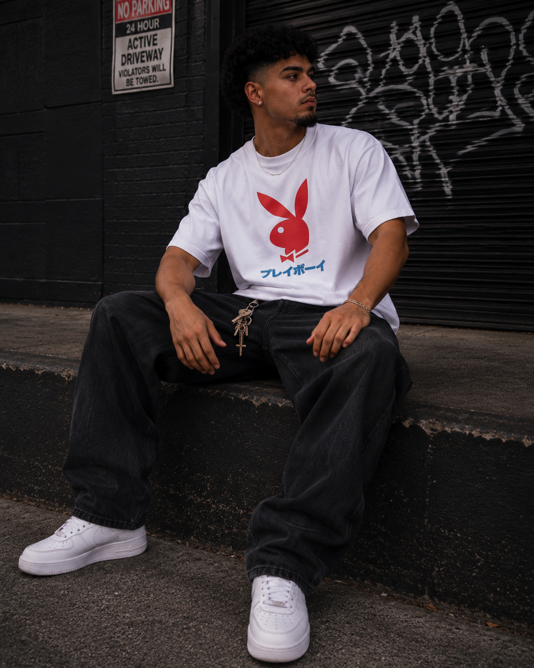 A man sitting on concrete steps in front of a graffiti-covered black metal shutter, wearing a white t-shirt with a red Playboy bunny logo and blue Japanese text, paired with dark baggy jeans and white sneakers. #Streetwearmen