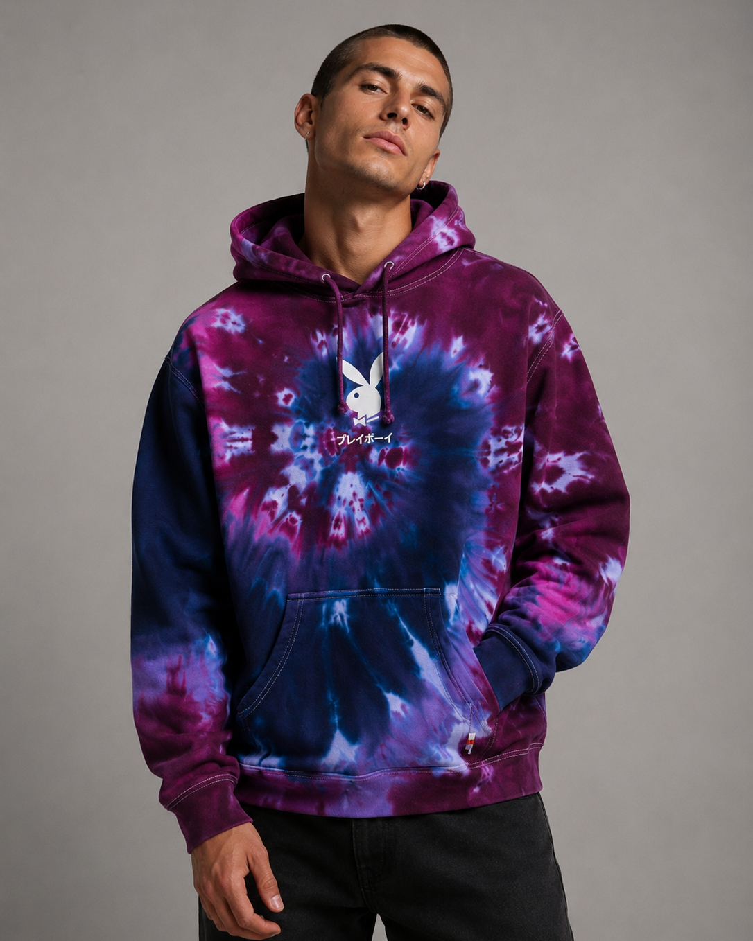 A man wearing a purple and blue tie-dye hoodie with a white Playboy bunny logo on the chest, posed against a solid gray studio background.
