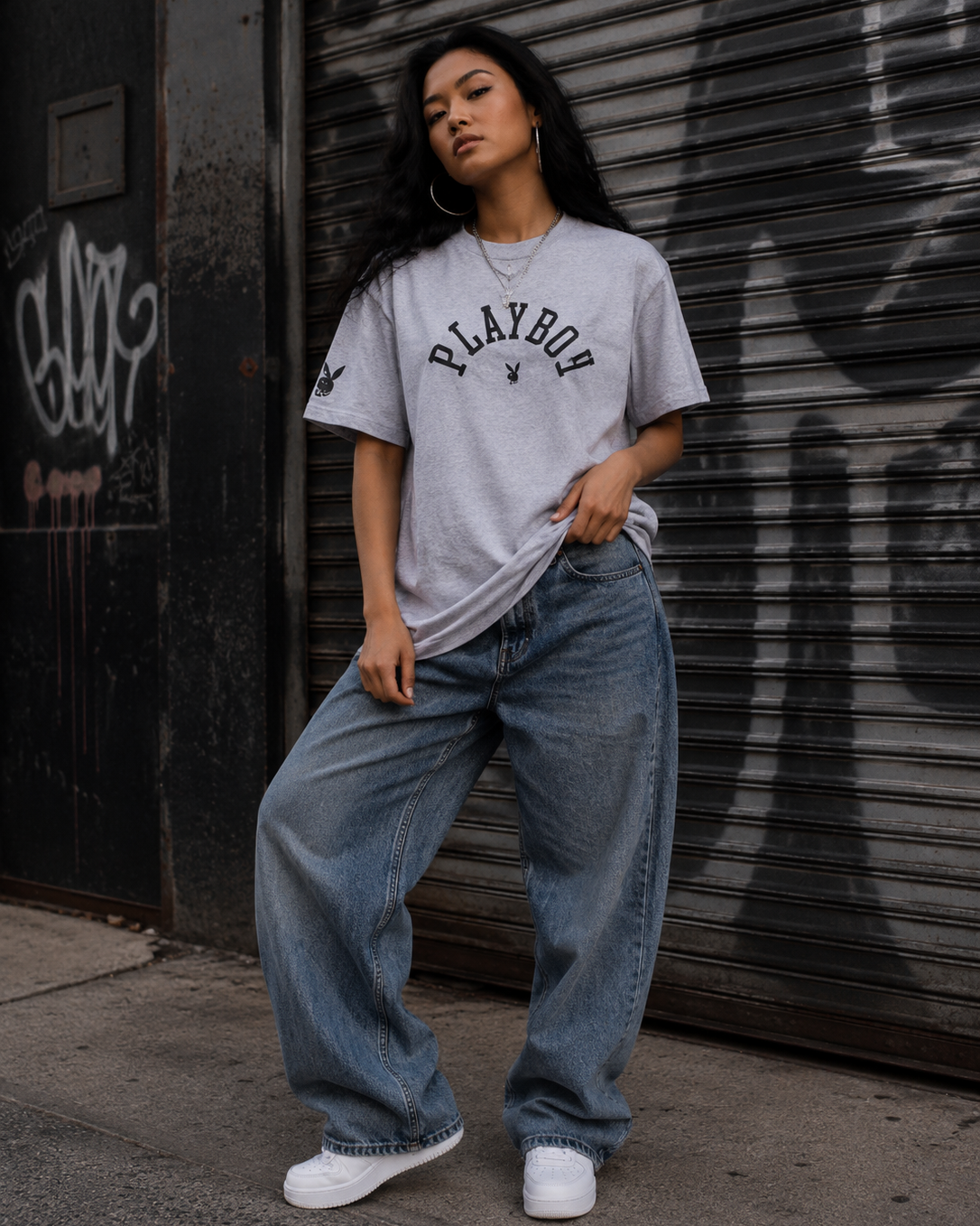 A woman stands in front of a closed metal storefront door wearing a light gray Playboy t-shirt, loose light-wash blue jeans, and white sneakers. #Streetwearwomen
