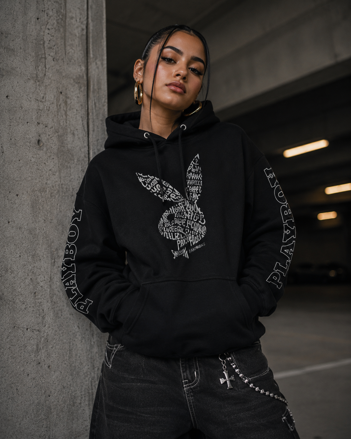 A woman wearing a black Playboy hoodie with a white graphic on the front and 'PLAYBOY' text down both sleeves, standing in a concrete parking structure. #Streetwearwomen