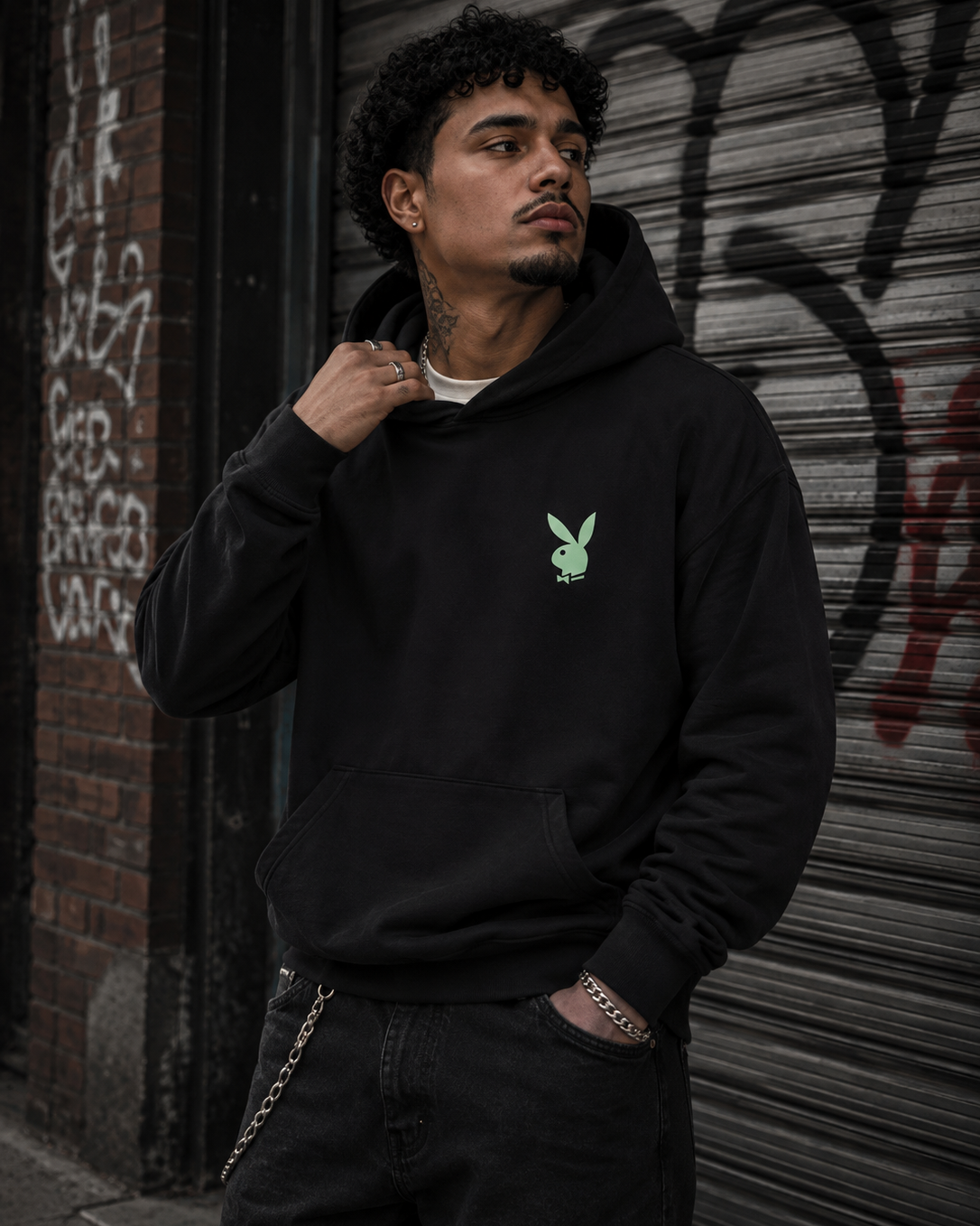 A man wearing a black hoodie with a small green Playboy bunny logo on the left chest, standing in front of a graffiti-covered metal shutter. #Streetwearmen