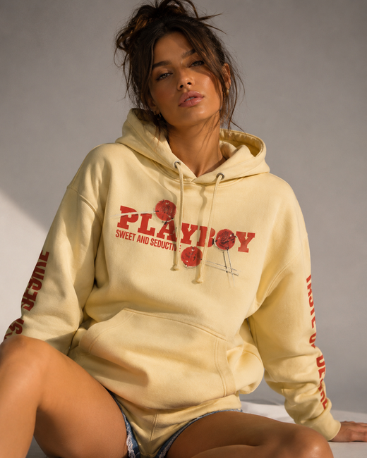 A woman wearing a pale yellow hoodie with a red 'Playboy Sweet and Sexy' graphic sits against a neutral gray background.