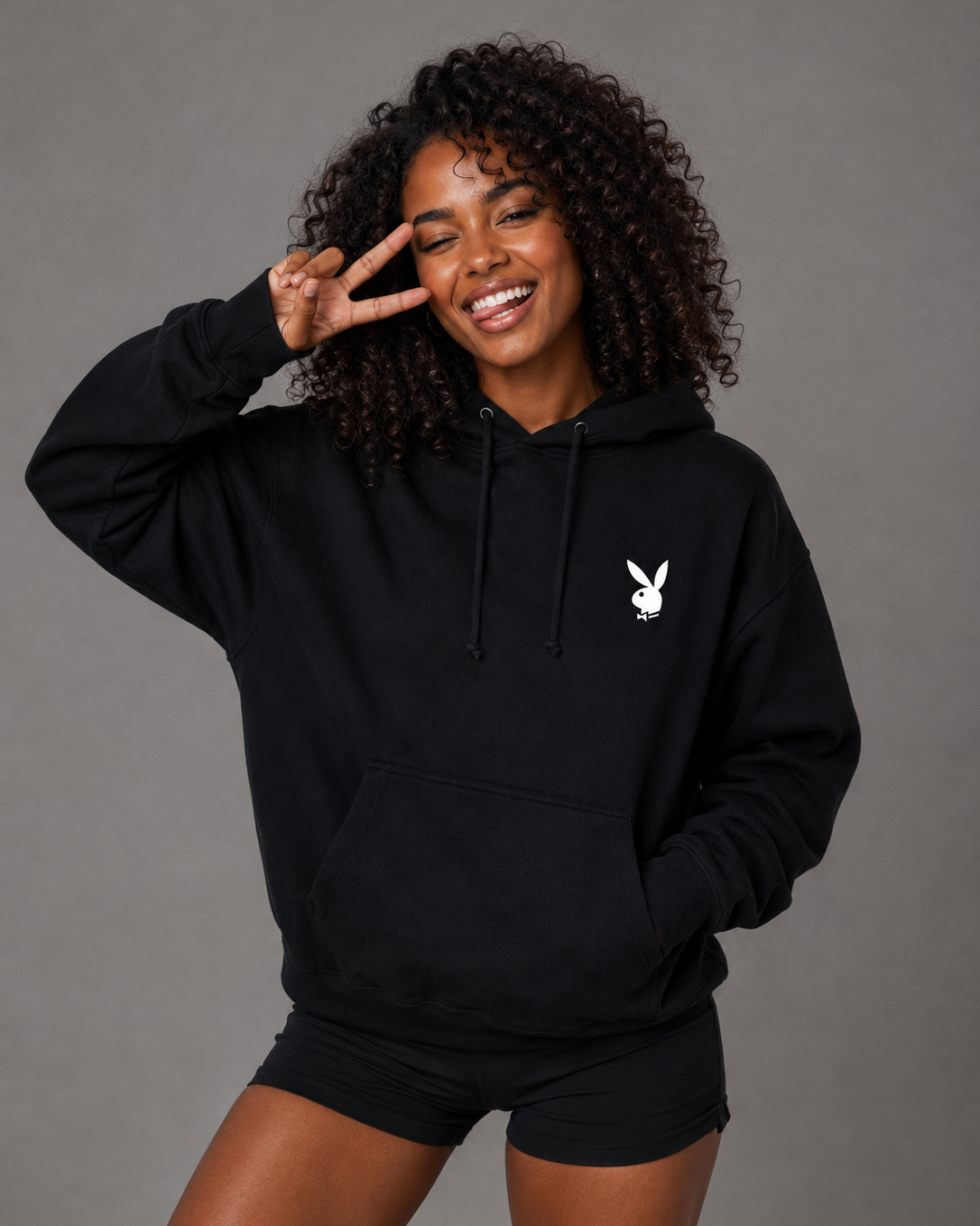 A woman with curly dark hair models a black pullover hoodie featuring a small white Playboy bunny logo on the left chest, paired with black athletic shorts, against a solid gray studio background.