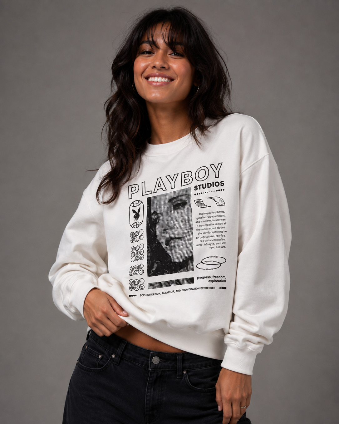 A woman wearing a white Playboy crewneck sweatshirt with a black graphic print and black jeans, posing against a solid gray studio background.