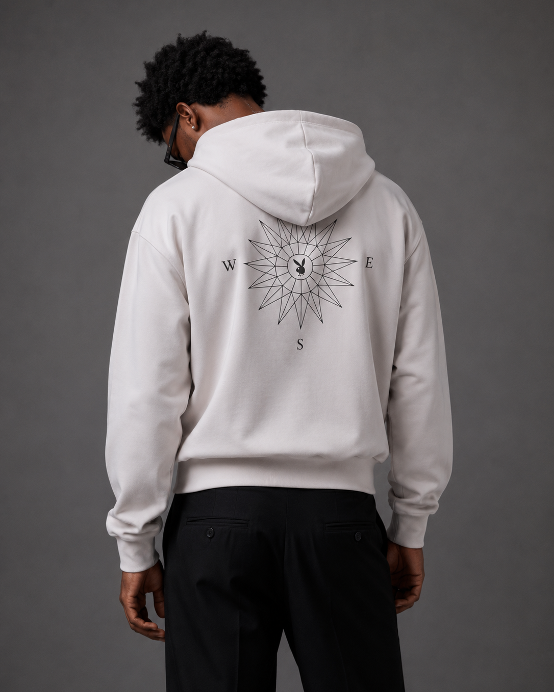 A man viewed from behind wearing a light gray hoodie with a detailed compass rose graphic printed on the back, paired with black pants, against a solid dark gray studio background.