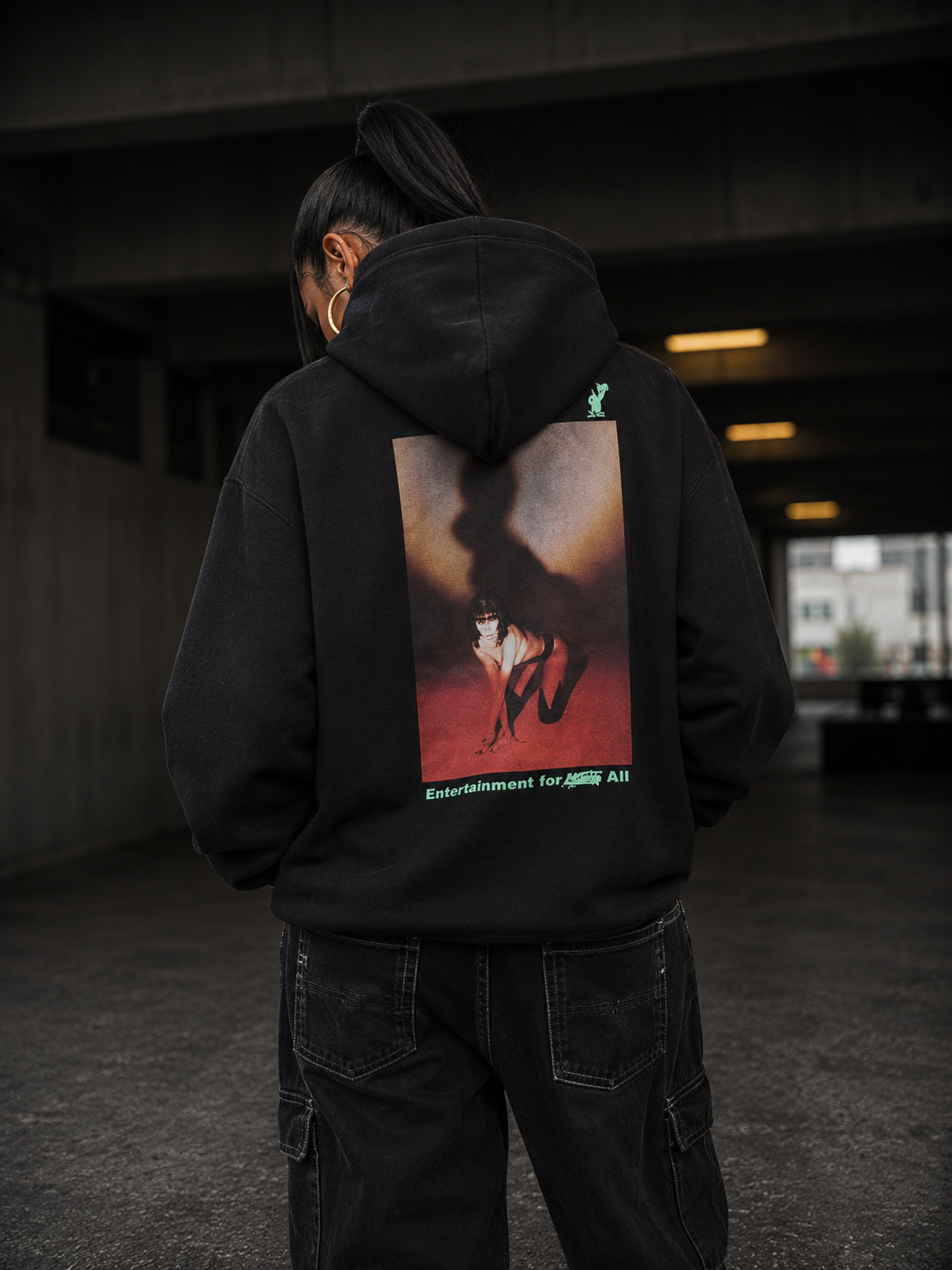 A person viewed from behind wearing a black hoodie with a large rectangular graphic print on the back, standing in a dim concrete tunnel or parking structure. #Streetwearwomen