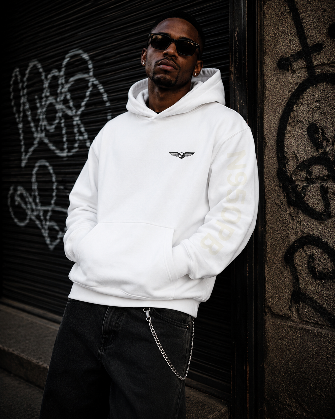 A man wearing a white pullover hoodie with a small black winged logo on the left chest and pale gold lettering down the right sleeve, paired with black jeans and sunglasses, leaning against a graffiti-covered dark metal shutter. #Streetwearmen