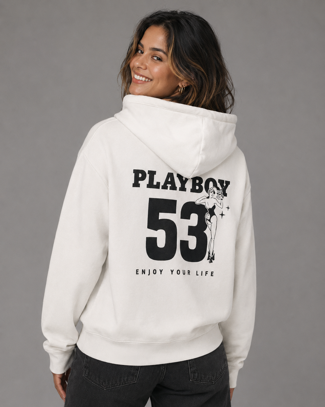 A woman wearing a white pullover hoodie with a large black PLAYBOY 53 graphic on the back, shown from behind against a plain gray studio background.
