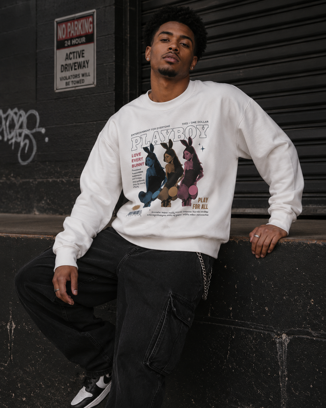 A man wearing a white Playboy crewneck sweatshirt with a vintage bunny graphic, paired with black cargo pants, leaning against a dark metal shutter in an urban setting. #Streetwearmen