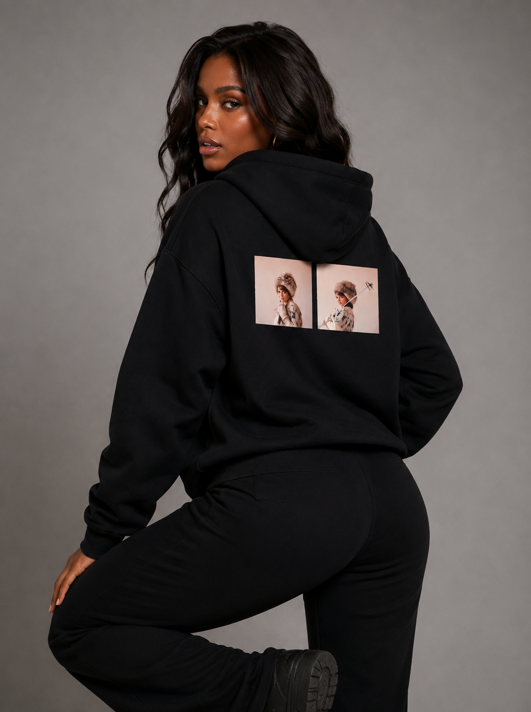 A woman posing from behind wearing a black hoodie with a graphic print on the back and matching black sweatpants, set against a solid gray studio background.