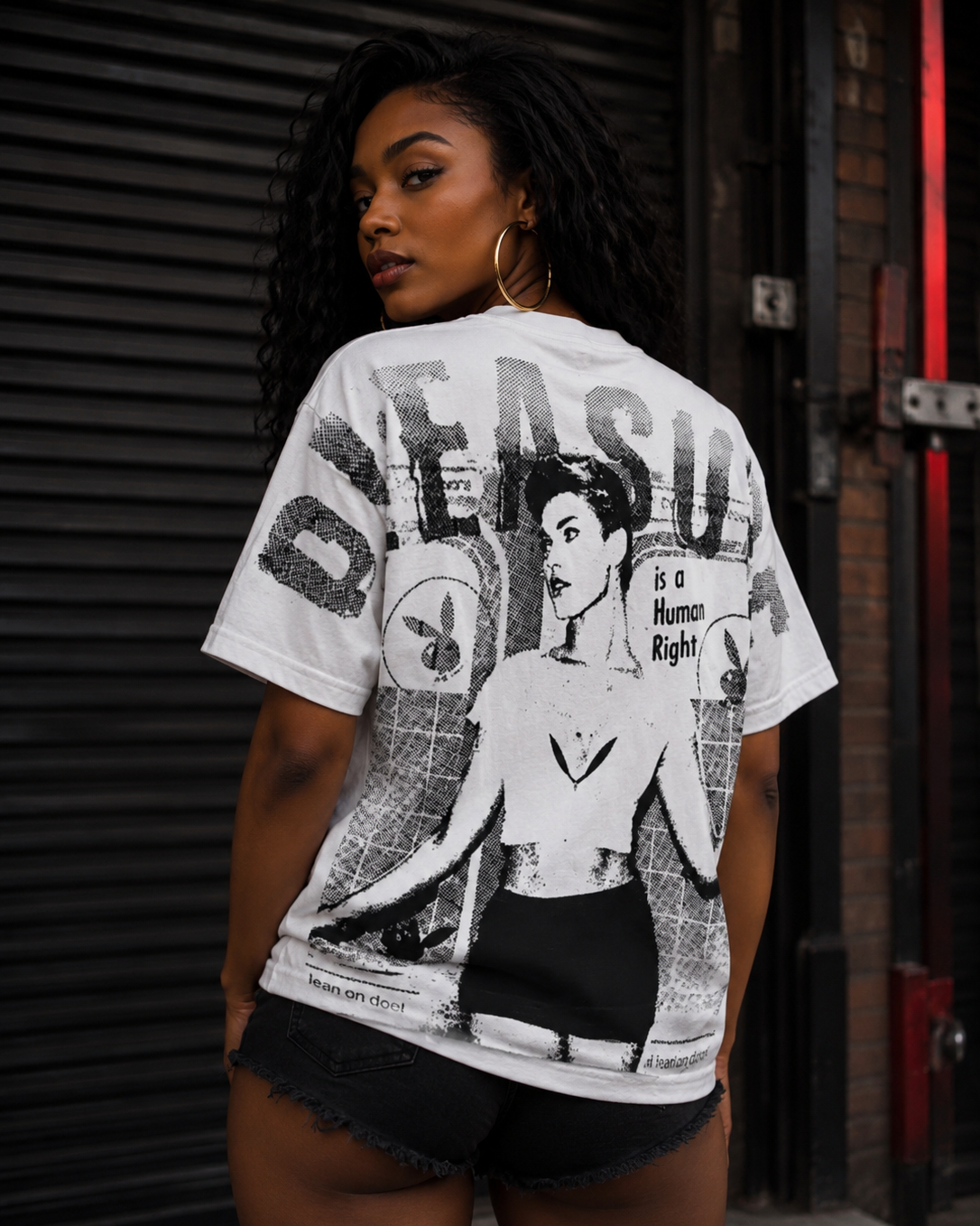 A woman wearing a white graphic t-shirt with a black and gray print of a woman and the word 'DESAVAGE', paired with black denim shorts, standing in front of a dark metal shutter. #Streetwearwomen