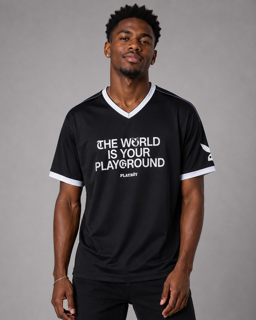 A man wearing a black v-neck t-shirt with white trim and white text that reads 'THE WORLD IS YOUR PLAYGROUND' and 'PLAYBOY' underneath, against a solid gray studio background.