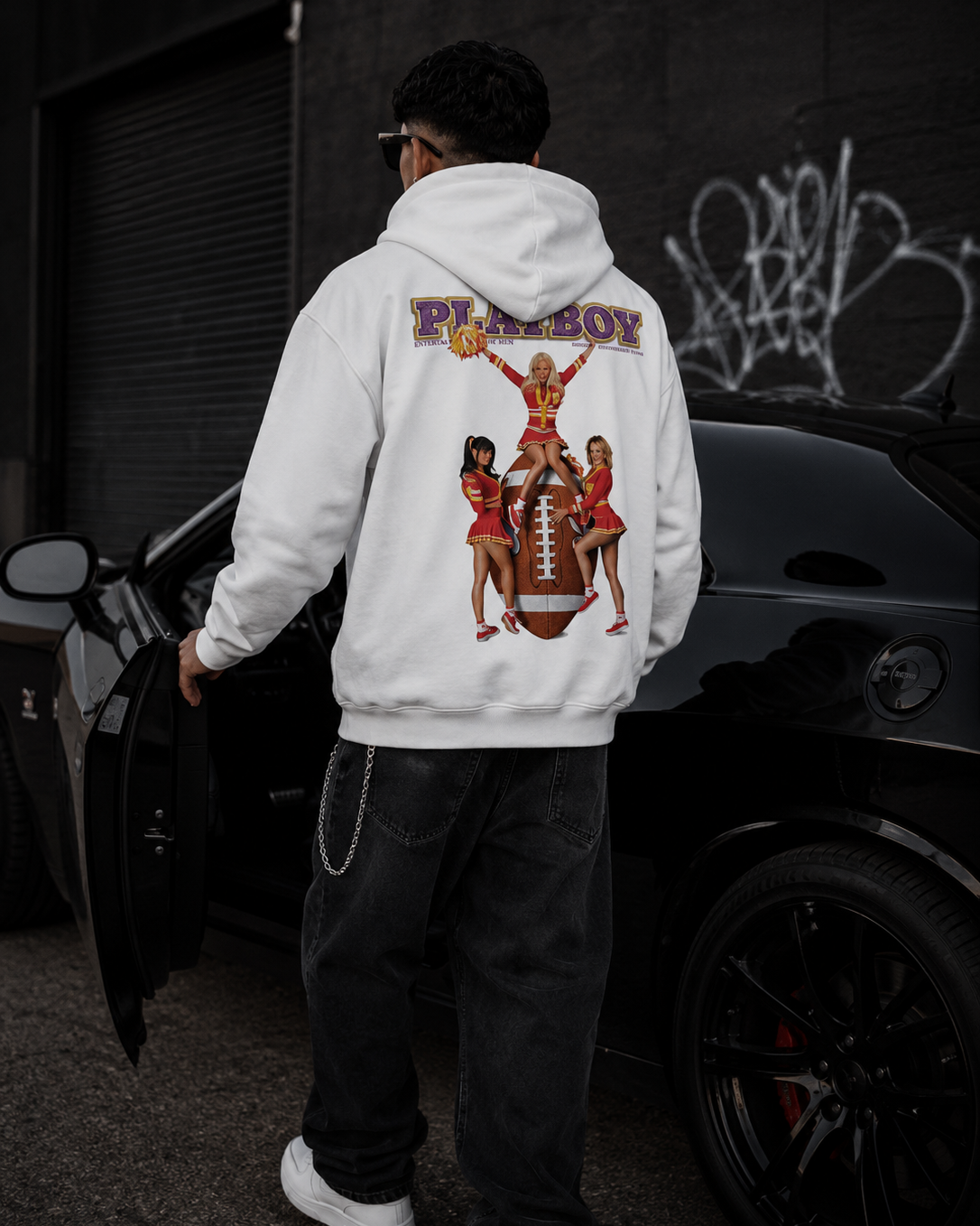 A man wearing a white Playboy hoodie with a large graphic print on the back, standing next to a black car in an urban setting. #Streetwearmen