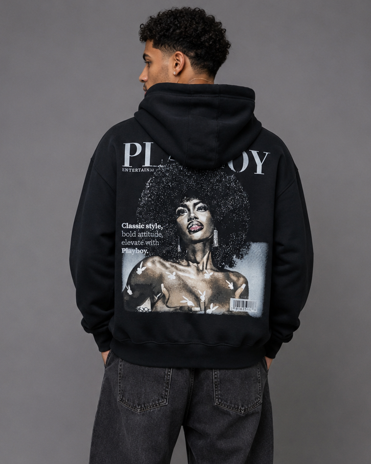 A man viewed from behind wearing a black hoodie with a large Playboy magazine cover graphic on the back, paired with dark gray jeans and white sneakers, against a solid gray studio background.