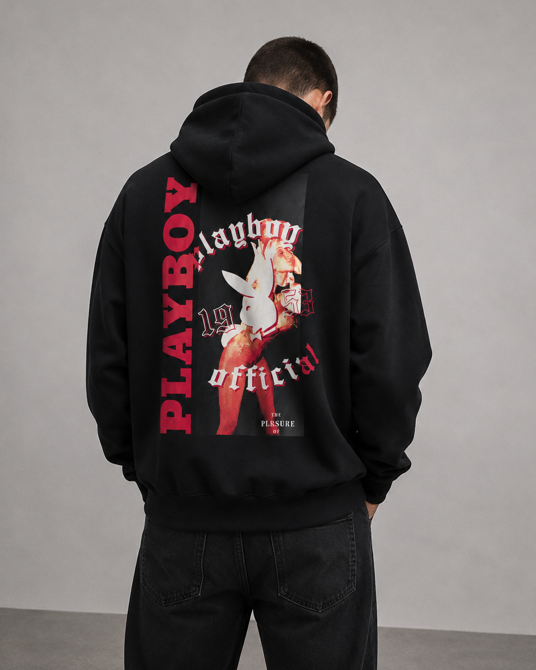 A man viewed from behind wearing a black hoodie with a large red and white Playboy back graphic print, paired with black jeans, against a neutral gray studio background.