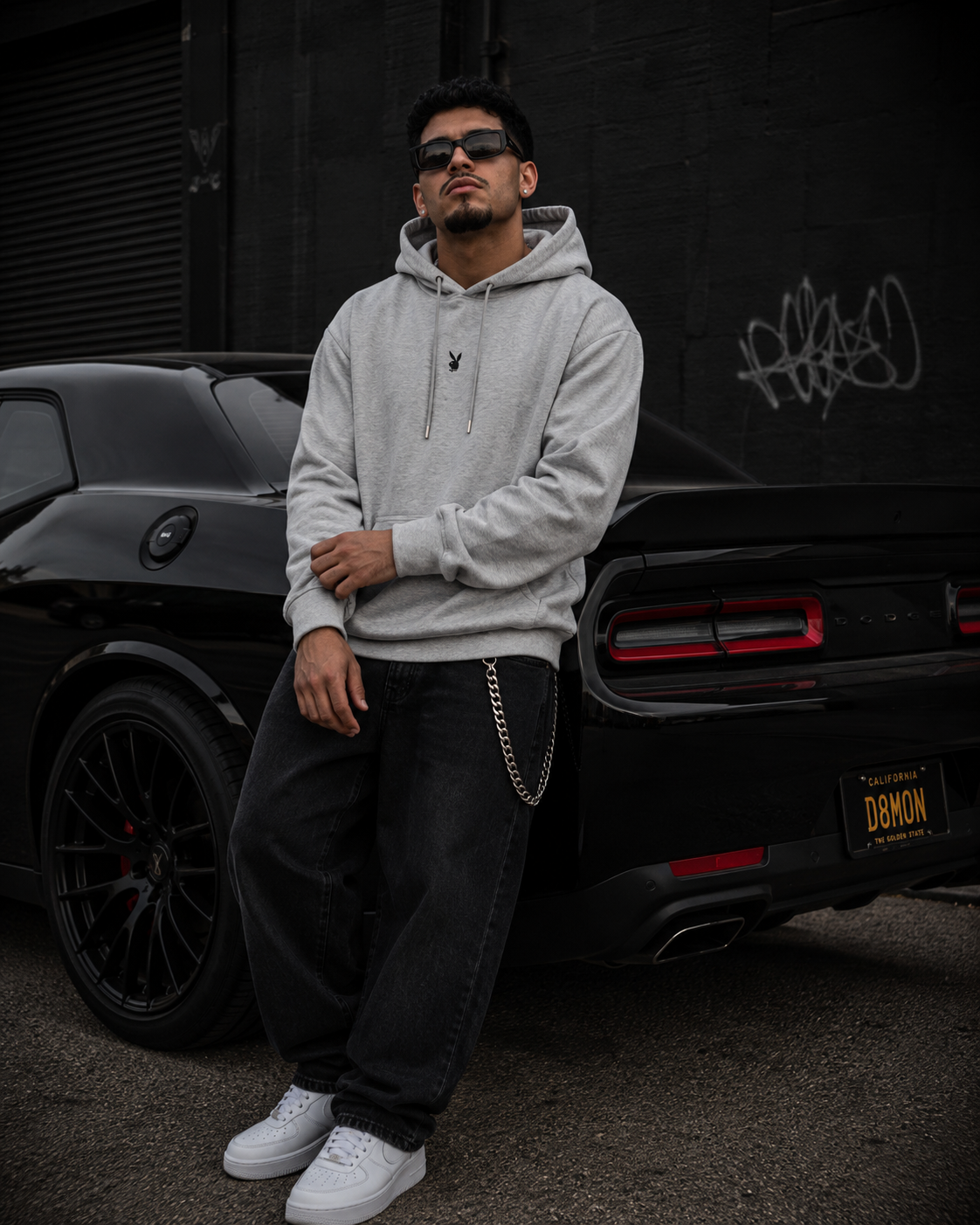 A man wearing a light gray hoodie, black jeans, and white sneakers stands next to a black car in front of a dark wall with graffiti. #Streetwearmen