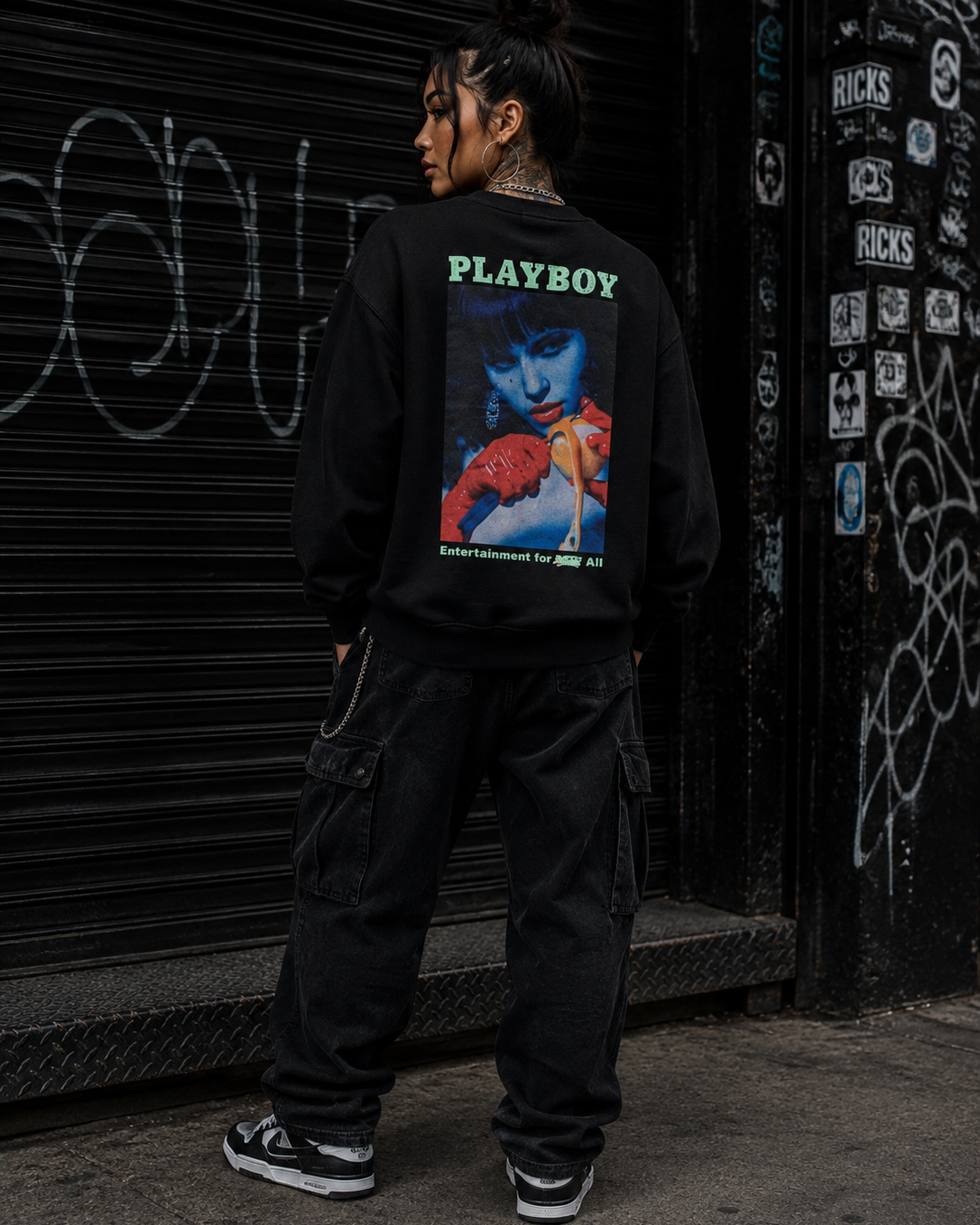 A person standing in front of a black metal shutter and graffiti-covered wall, wearing a black sweatshirt with a large back graphic that reads PLAYBOY and shows a colorful portrait print, paired with black cargo pants and sneakers. #Streetwearwomen