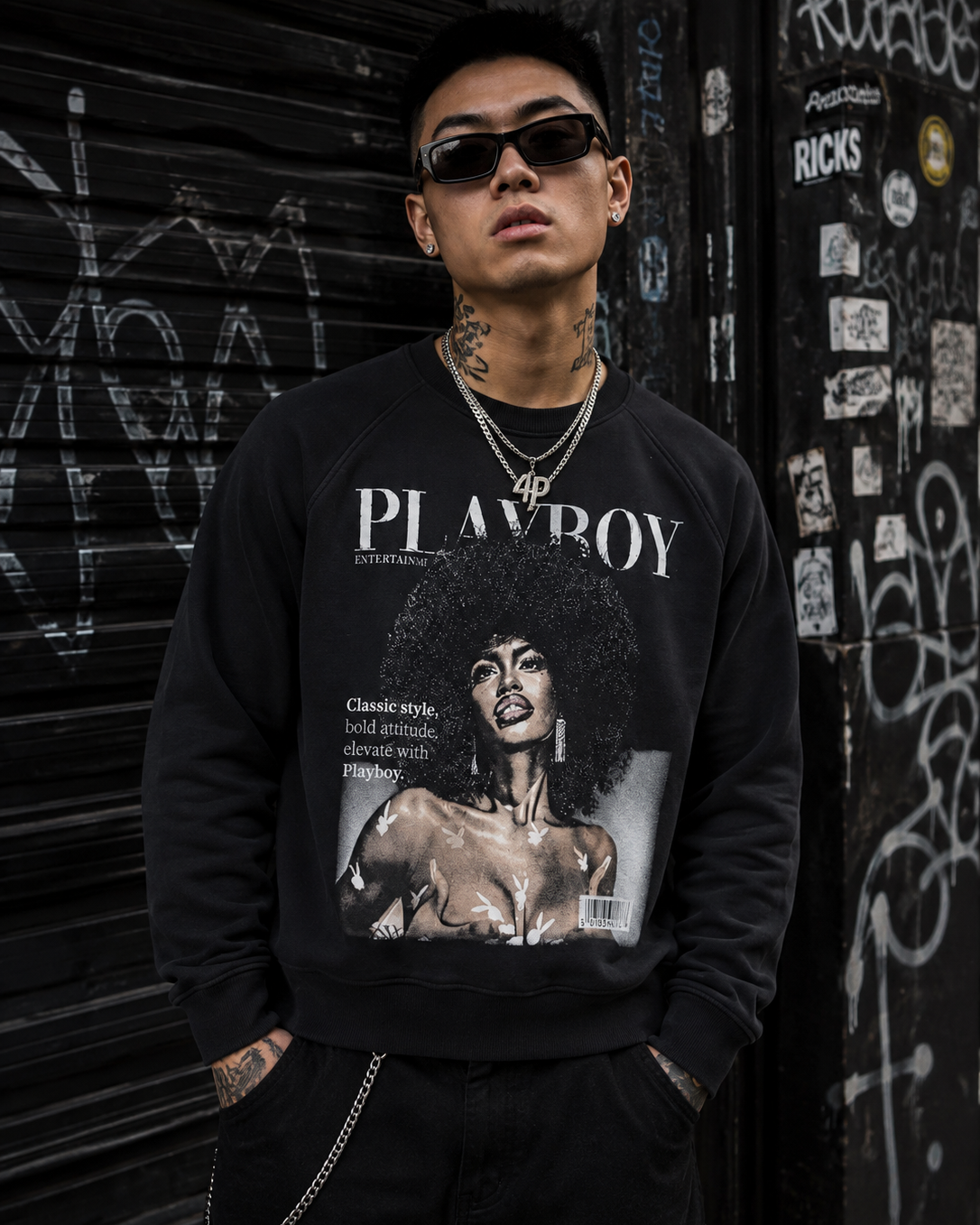 A man wearing a black Playboy crewneck sweatshirt with a large vintage-style graphic print on the front, standing in front of a graffiti-covered metal shutter. #Streetwearmen