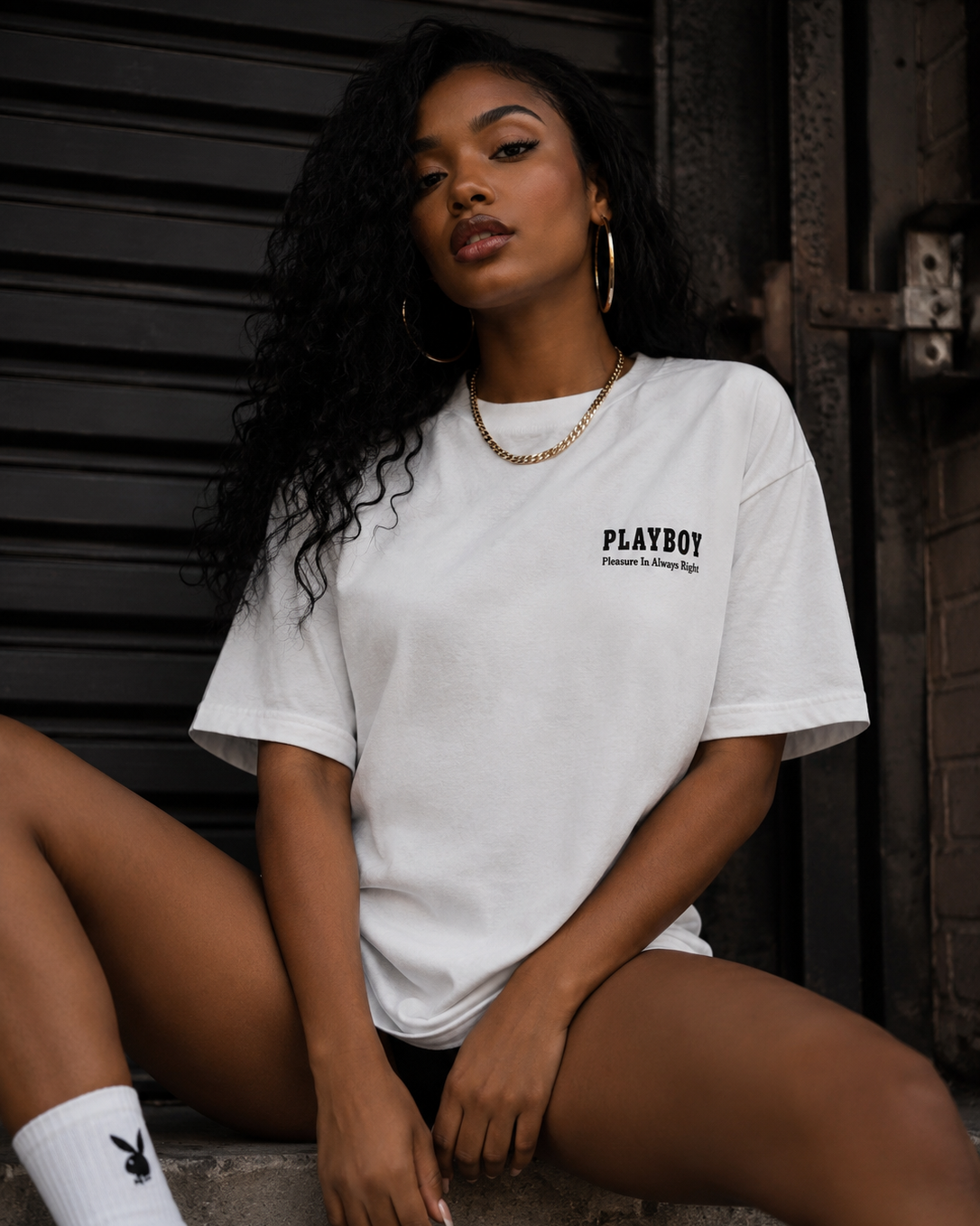 A woman wearing a white PLAYBOY t-shirt with black text on the left chest, sitting against a dark metal shutter background. #Streetwearwomen