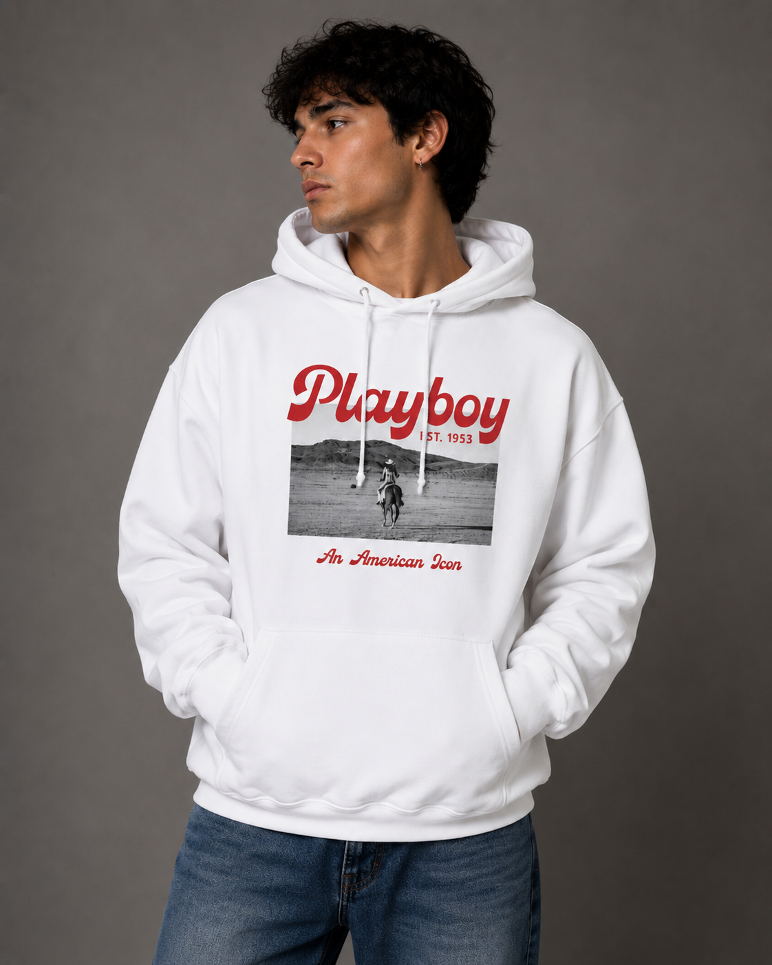A man wearing a white Playboy hoodie with a red and black graphic print on the front, paired with blue jeans, standing against a solid gray studio background.