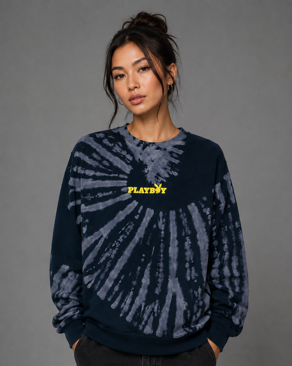 A woman modeling a dark navy blue tie-dye sweatshirt with a yellow 'PLAYBOY' logo on the chest, paired with black pants, against a solid gray studio background.