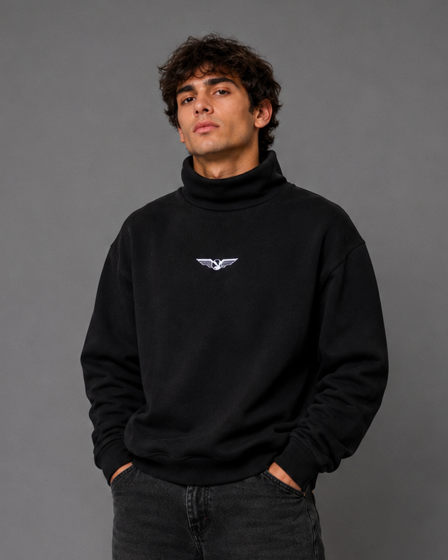 A man modeling a black turtleneck sweatshirt with a small white wing logo on the left chest, paired with dark gray jeans and black sneakers, standing against a solid gray studio background.