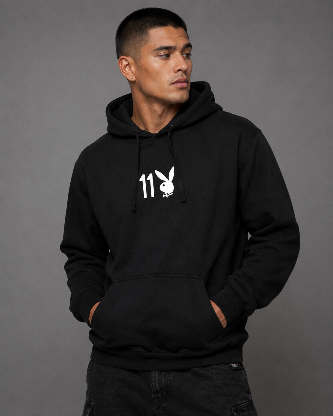 A man wearing a black pullover hoodie with a white Playboy bunny and '11' graphic on the chest, posed against a solid gray studio background.