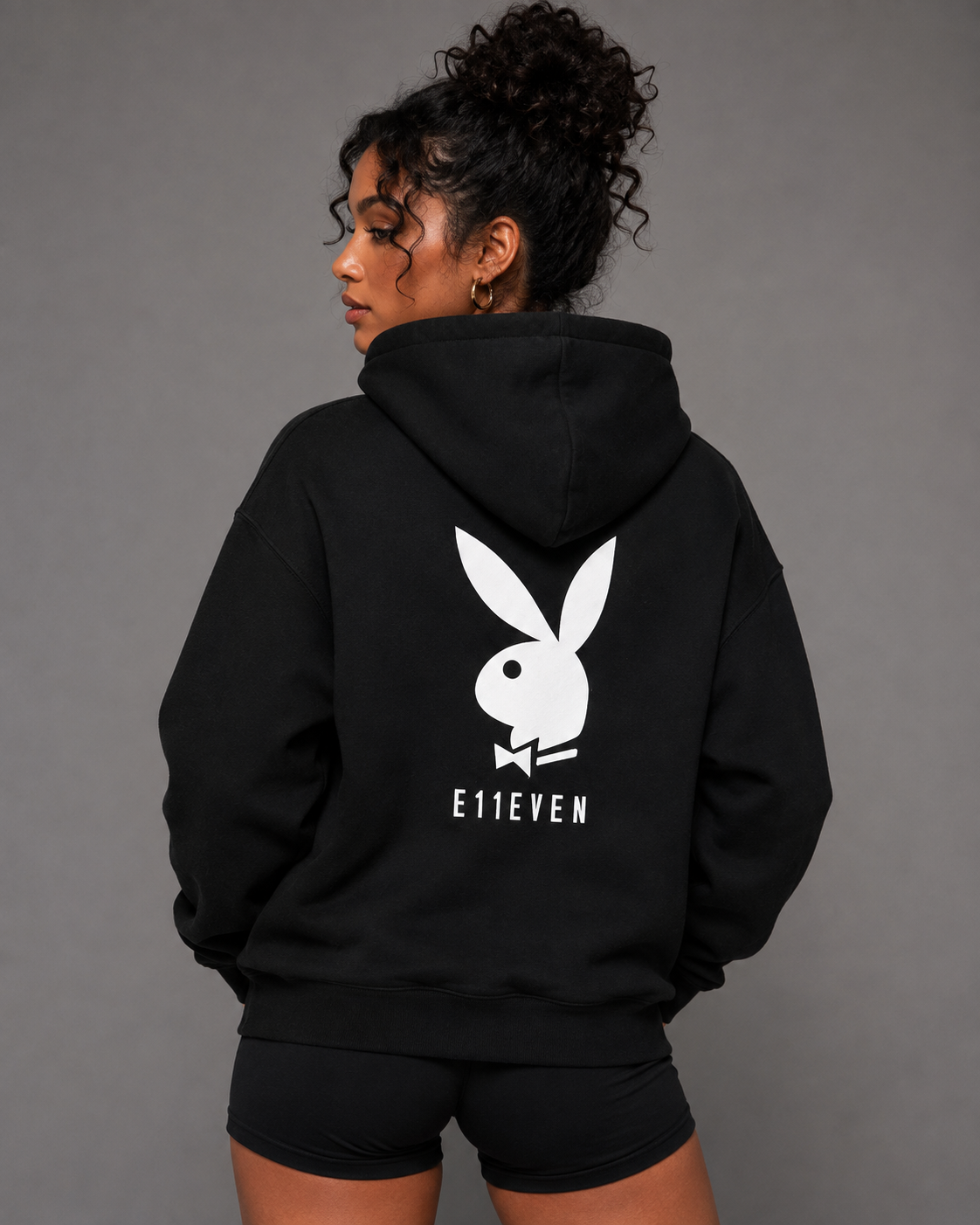 A woman shown from the back wearing a black hoodie with a large white Playboy bunny logo and the text 'E11EVEN' printed on the back, paired with black shorts against a solid gray studio background.