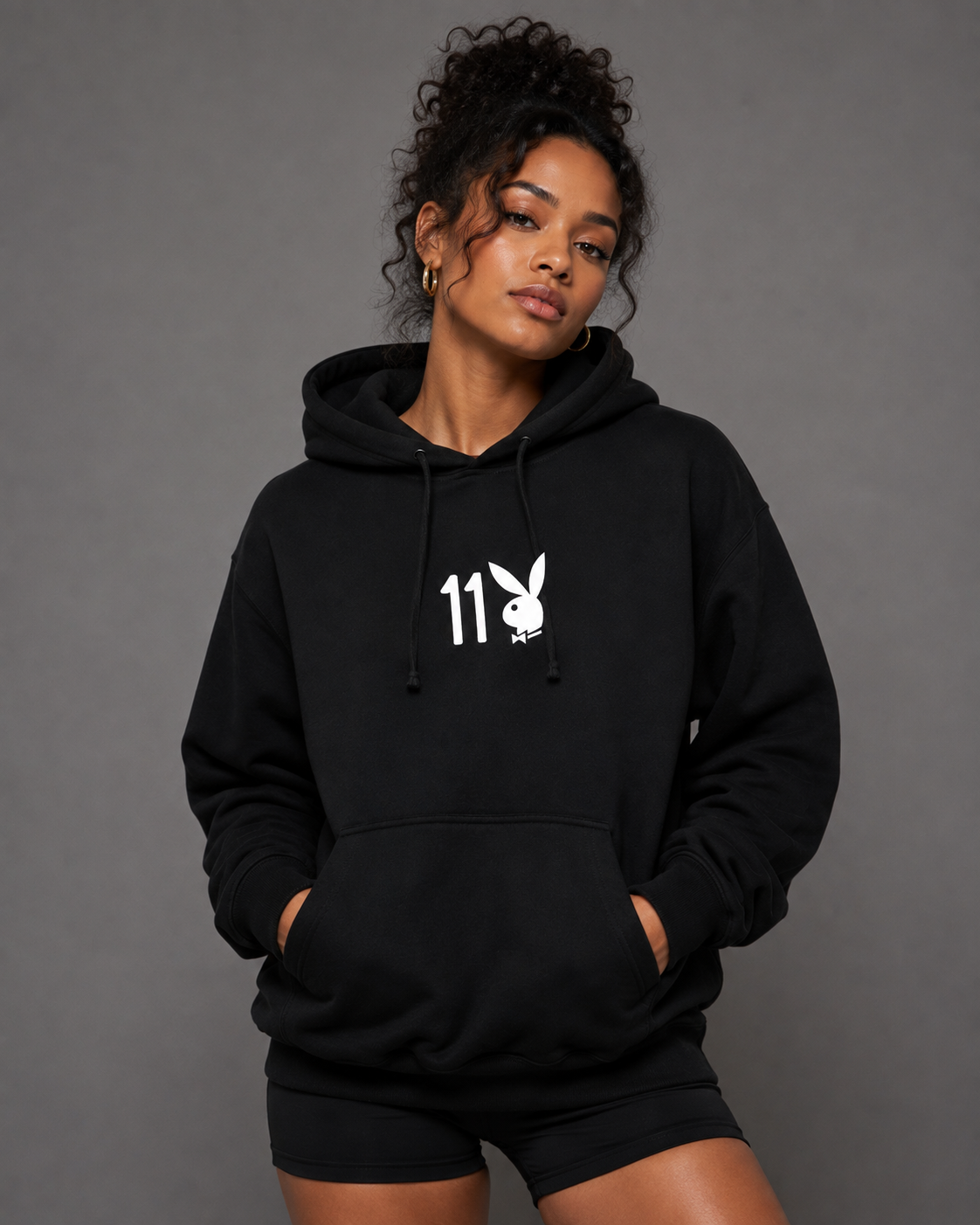 A woman modeling a black pullover hoodie with a white graphic on the chest featuring the number 11 and the Playboy bunny logo, paired with black athletic shorts against a solid gray studio background.