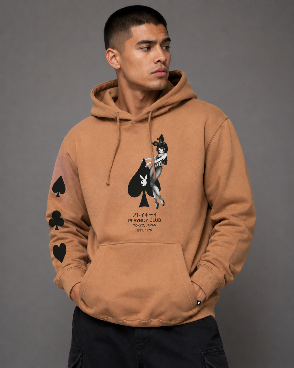 A man wearing a tan hoodie with a black and white graphic of a playing card character on the chest, along with black spade and heart graphics on the sleeves. The text on the chest reads 'PLAYER PARKER CLUB'.