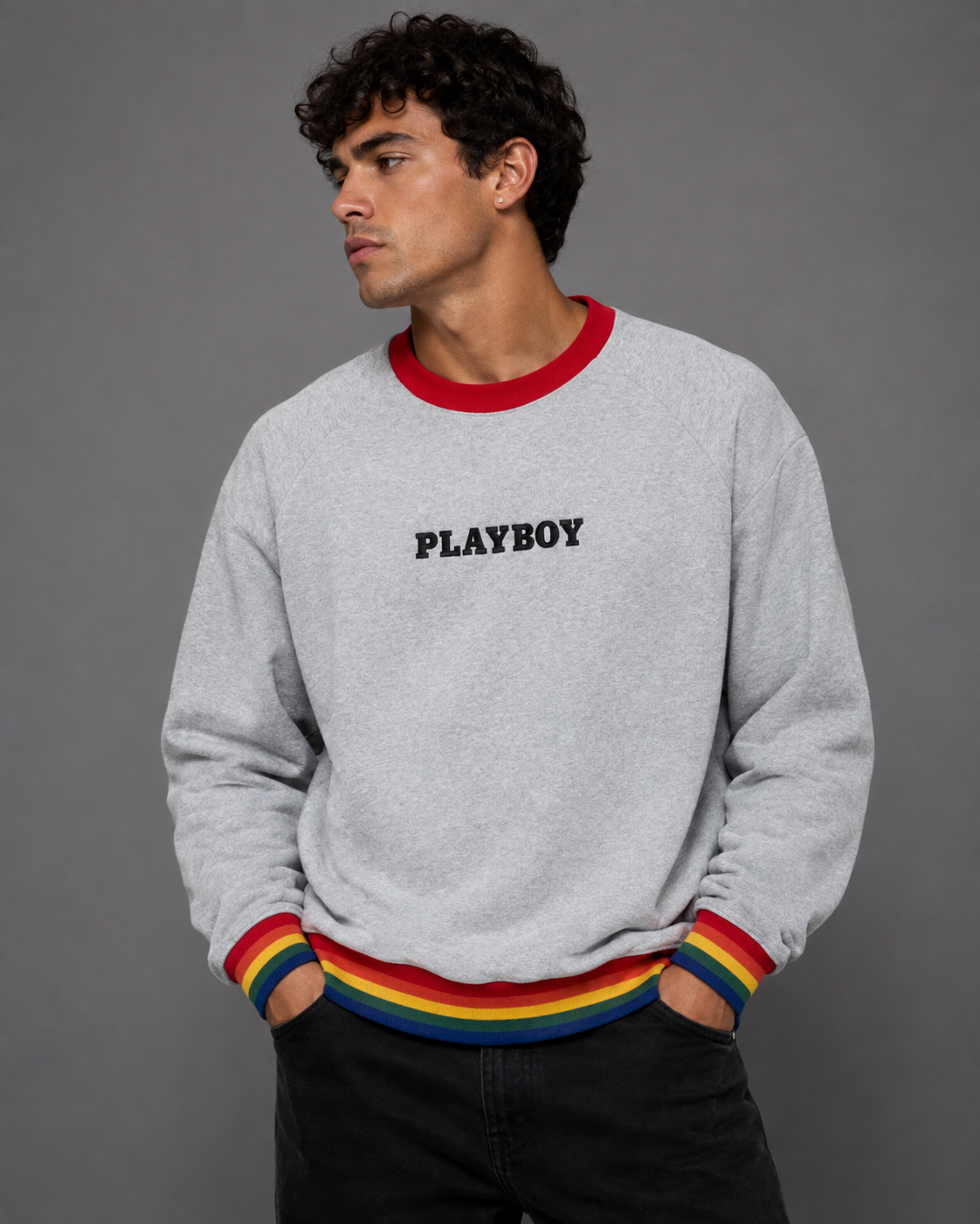 A man wearing a light gray heather crewneck sweatshirt with a red collar, rainbow-striped cuffs and hem, and the black Playboy logo on the chest, posed against a solid gray studio background.