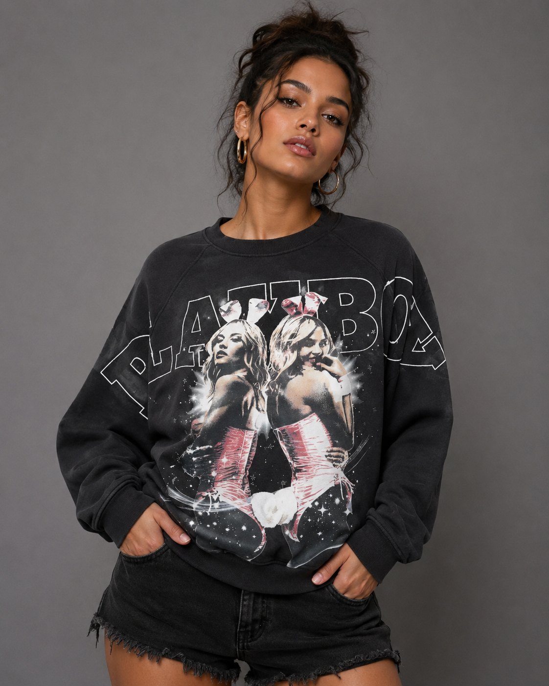 A woman modeling a black graphic sweatshirt featuring a large white 'PLATYBO' text and a photo print of two women in bunny suits, paired with black denim shorts against a solid gray studio background.