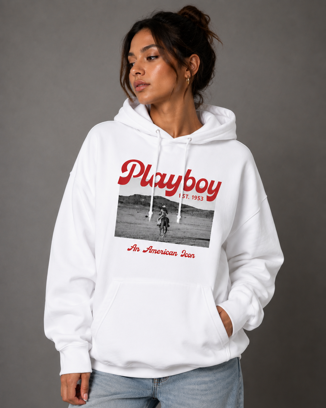 A woman wearing a white pullover hoodie with a red and black 'Playboy' graphic on the front, paired with light blue jeans, photographed against a neutral gray studio background.