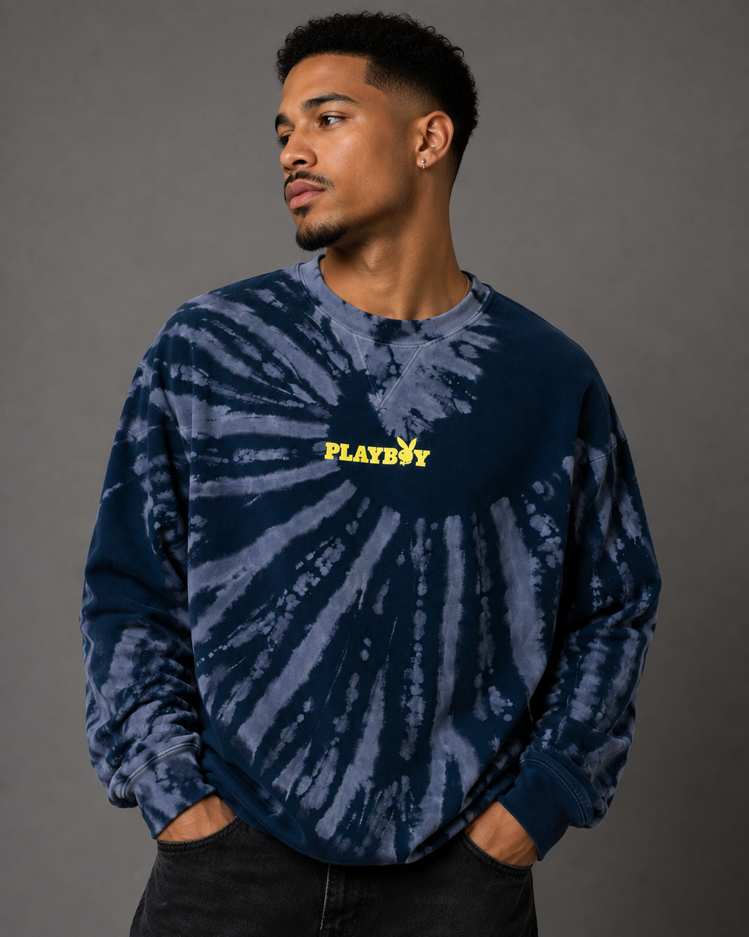 A man wearing a blue and navy tie-dye sweatshirt with a yellow PLAYBOY logo on the chest, paired with black jeans, against a dark gray studio background.