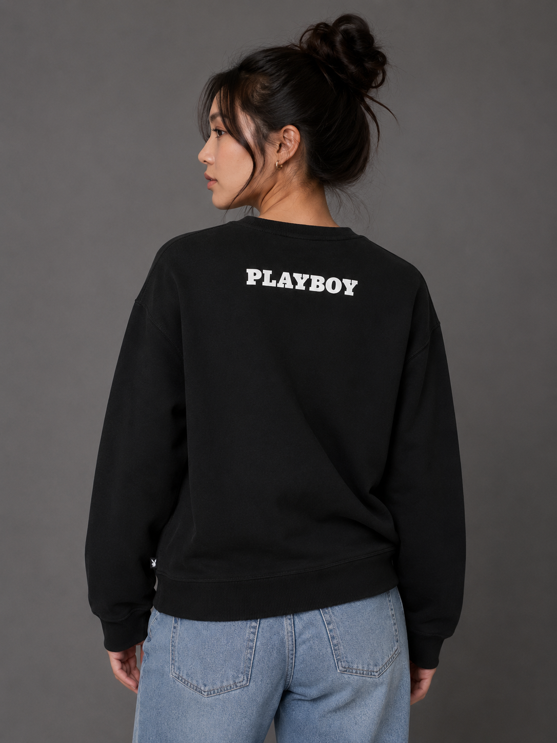 A woman shown from the back wearing a black sweatshirt with 'PLAYBOY' printed in white across the upper back, paired with light blue jeans, against a dark gray studio background.