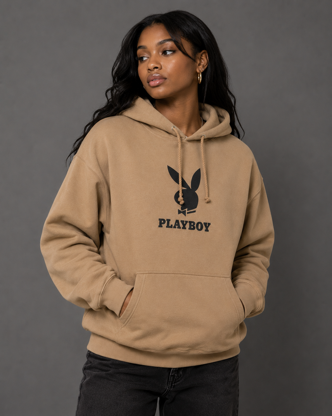 A woman wearing a tan oversized hoodie with a black Playboy bunny logo and the word 'PLAYBOY' printed on the chest, posed against a dark gray studio background.